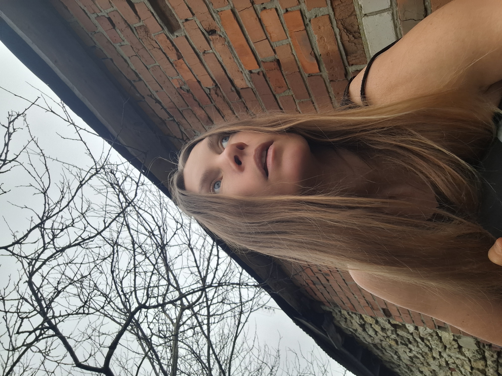 A low-angle, color photograph depicts a woman with long, straight, light brown hair looking upwards. Her eyes are blue. The background includes bare tree branches against a gray, overcast sky on the left. On the right, there is a brick wall with apparent damage. The picture's perspective suggests the subject may be lying down or leaning far back while taking the photo. Based on the query, the location is assumed to be Khlevnoye, Russia.