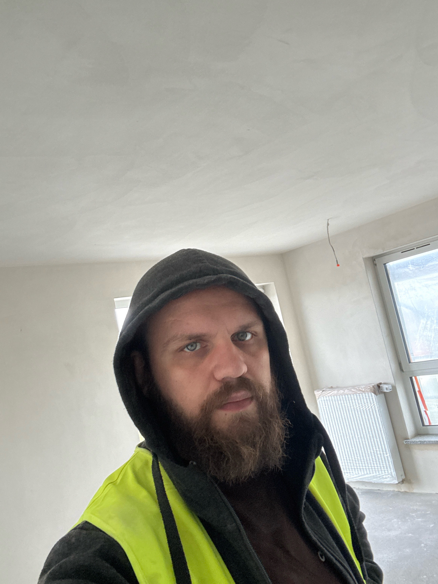 A man with a beard and blue eyes is taking a selfie in what appears to be an unfinished apartment in Nowa Sól, Poland. He is wearing a dark hooded sweatshirt and a bright yellow safety vest over it. The room is sparsely furnished, with bare walls and a concrete floor. A large window with a view of scaffolding can be seen to the right, and a radiator is installed beneath it. The ceiling is textured and a single electrical wire hangs from it.
