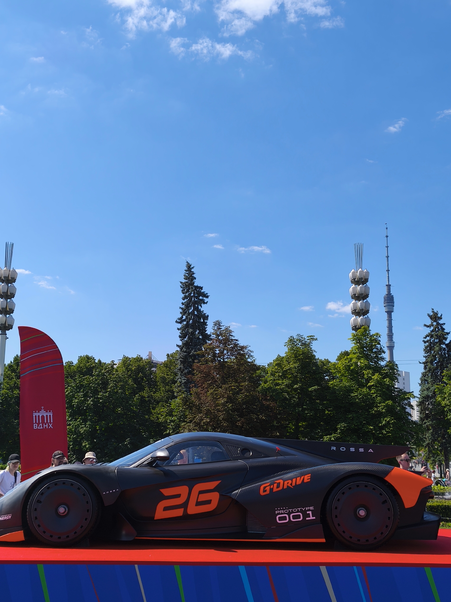 On a sunny day in Moscow, Russia, at the Exhibition of Achievements of National Economy (VDNKh), a sleek black sports car with orange accents is displayed on a red platform. The car features the logos "G-Drive" and "ROSSA" along with the number "26" prominently displayed on its side, and "ROSSA PROTOTYPE 001" on the rear fender. In the background, lush green trees form a backdrop to the iconic Ostankino Tower, a tall television tower, and another distinctive tower structure with multiple spherical lights. Several people are visible, some standing near the car and others in the distance, observing the scene. The sky is a clear blue with a few scattered white clouds.
