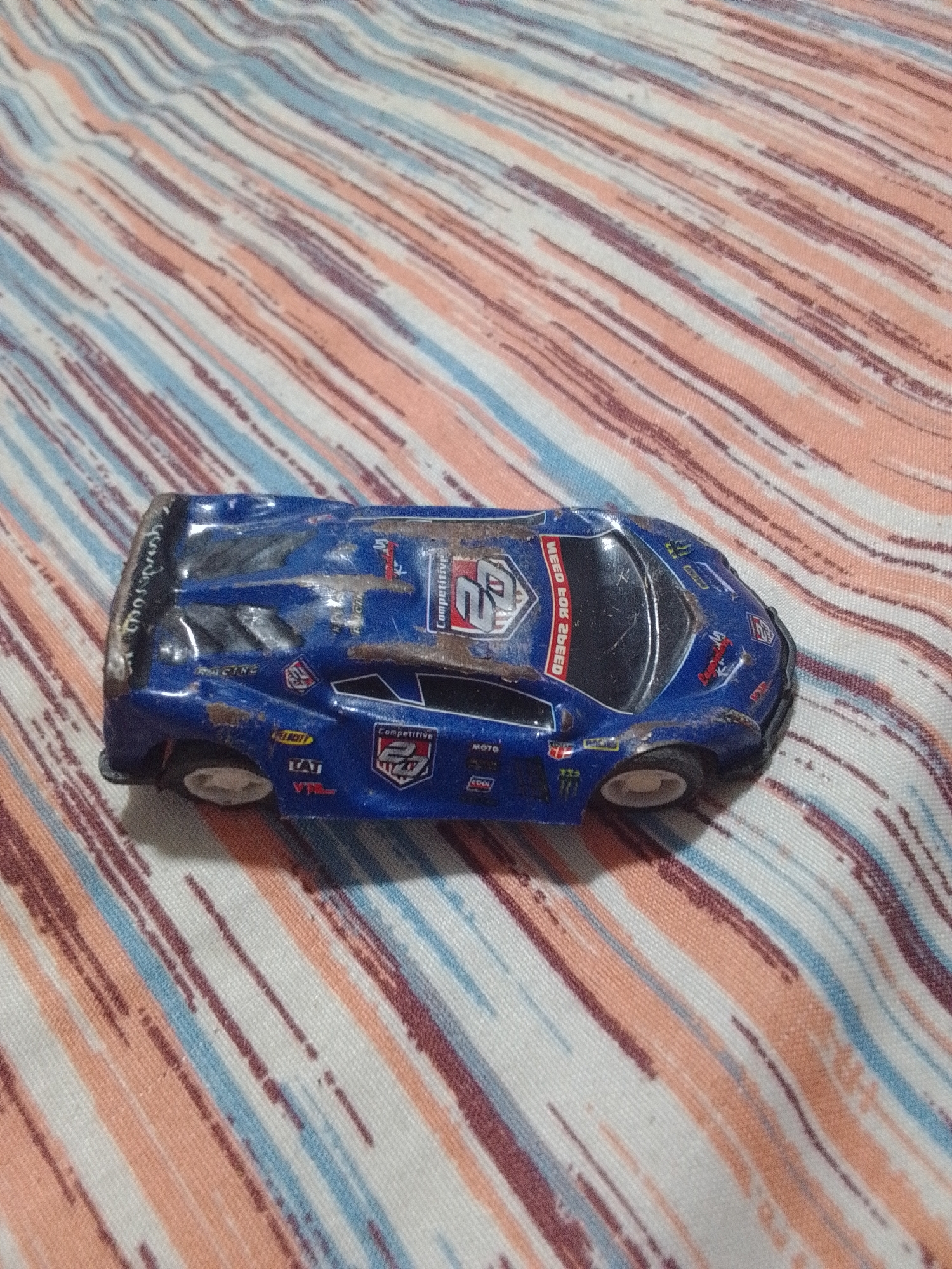 A blue toy race car, possibly a Lamborghini model, with racing decals and the text "NEED FOR SPEED" printed vertically on its side, is positioned on a striped fabric surface. The fabric has diagonal stripes in shades of orange, white, and blue-gray. The toy car appears worn, with scuff marks and chipped paint. The lighting suggests an indoor environment, and there are no people or specific urban elements visible that would indicate the location in La Tagua, Venezuela. The image is a close-up of the toy car and the fabric it rests on.