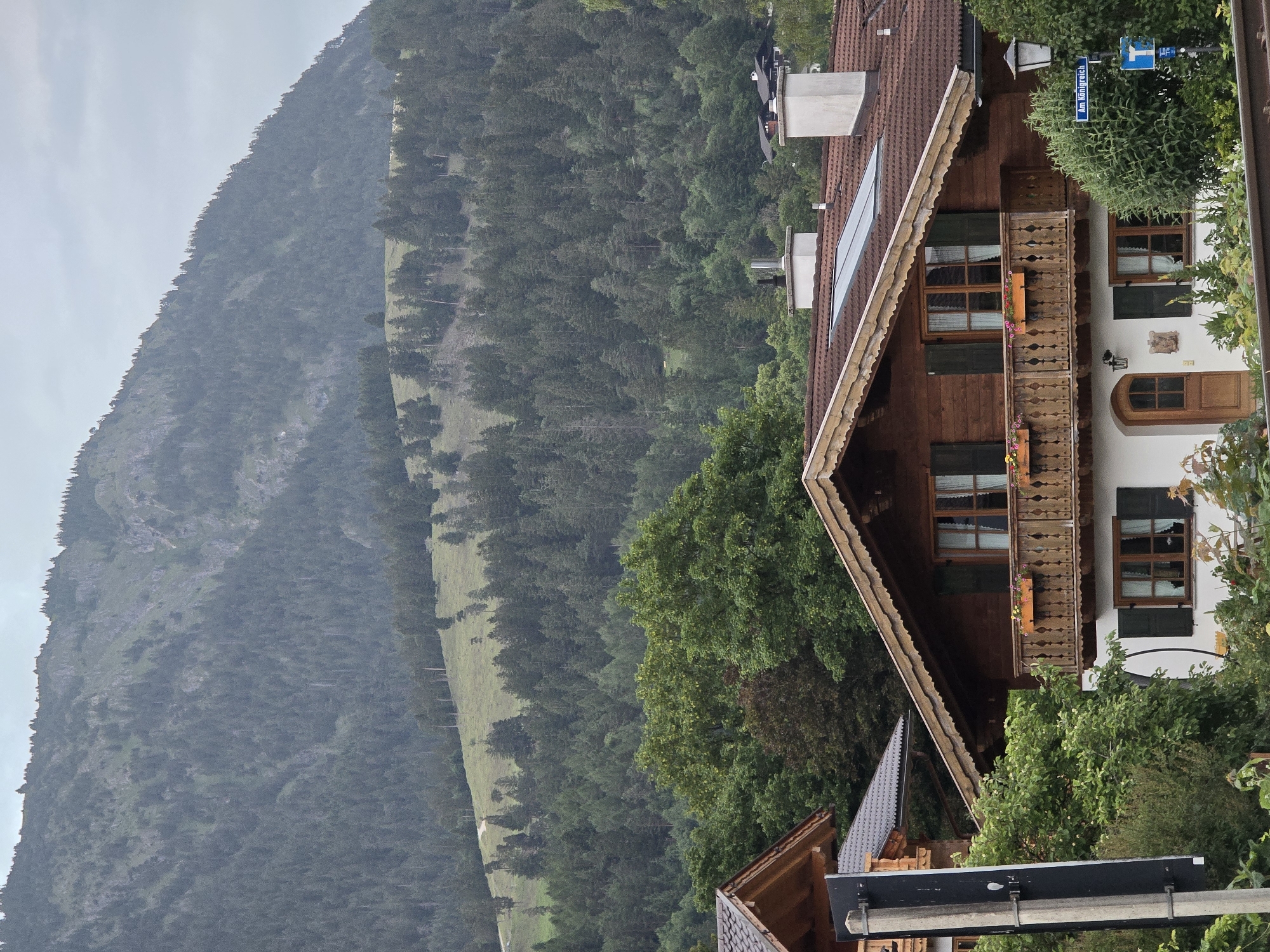 The image depicts a view of a traditional Bavarian house in Garmisch-Partenkirchen, Germany, with a densely forested mountain slope in the background. The house is constructed of wood with a brown tiled roof. It features wooden balconies with decorative railings and window boxes with flowers. Multiple windows with dark green shutters are visible. Two white chimneys protrude from the roof. A street sign reading "Am Königreisch" is attached to a lamppost on the right side of the image. The mountain behind the house is covered with evergreen trees, and the sky is overcast. The foreground shows some vegetation and what appears to be train tracks.