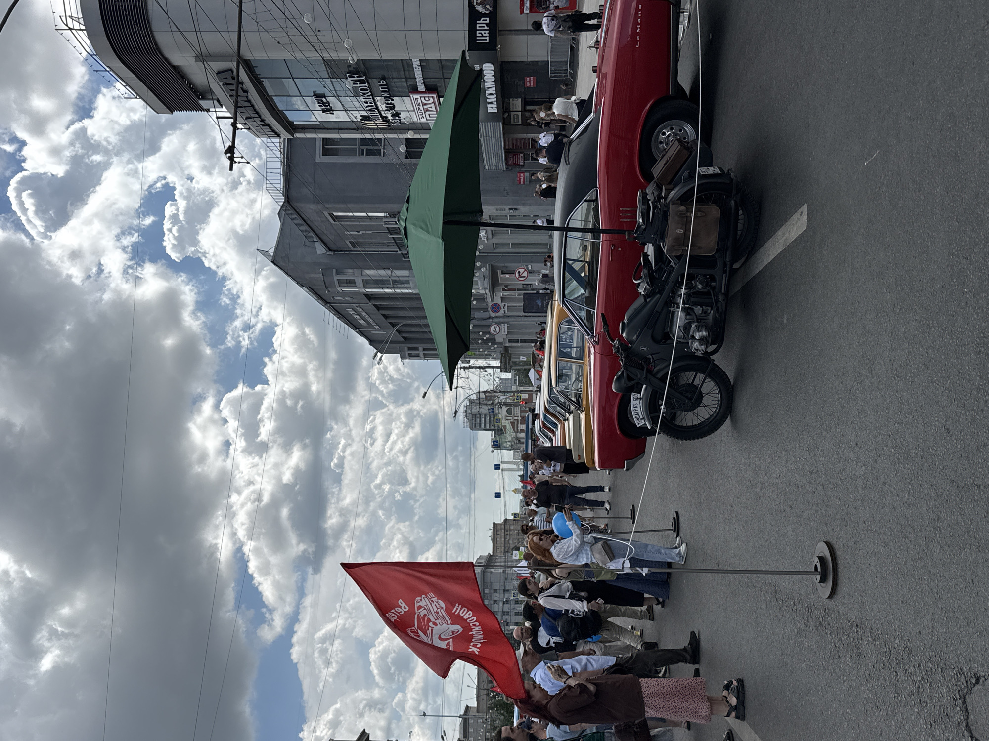 The image shows a street event, likely an exhibition of vintage cars and motorcycles, in Novosibirsk, Russia. A red flag with a white emblem and text in Russian, possibly related to a historical event or organization, is prominently displayed. Several vintage cars, including a red coupe, are parked along the street, and a motorcycle is positioned in the foreground. People are gathered on the sidewalk and street, observing the vehicles and the event. Buildings line the street, with some featuring signage in Russian. The sky is partly cloudy, indicating an outdoor event. The overall scene suggests a festive atmosphere.