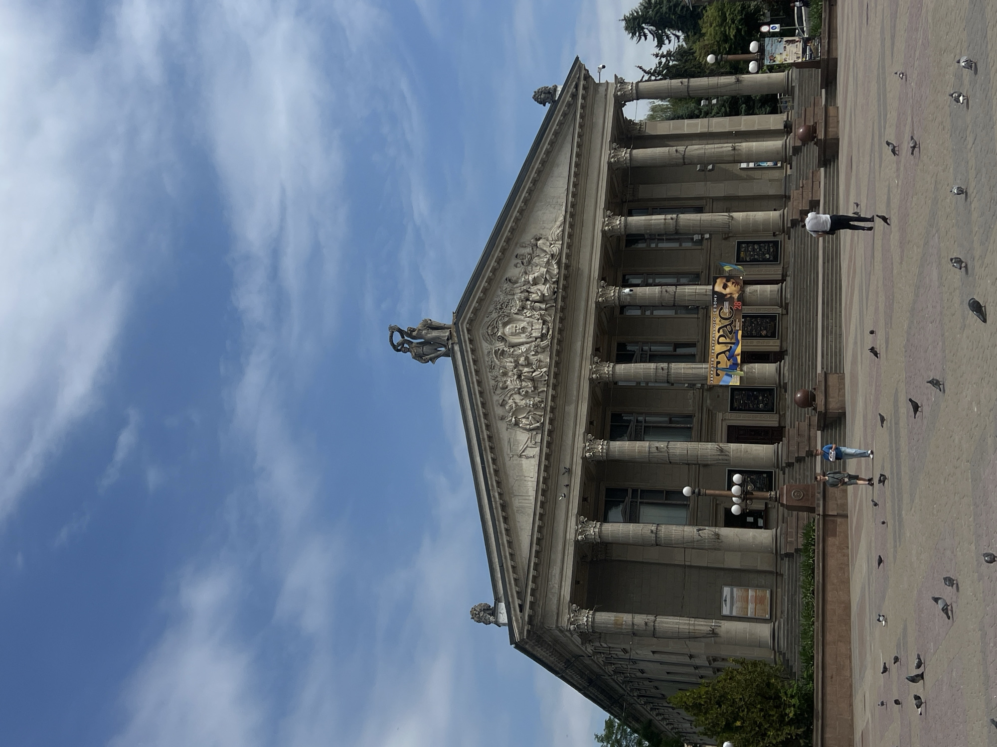 This is a photograph of the Ternopil Regional Drama Theatre, also known as the Ternopil Academic Ukrainian Drama Theatre, in Ternopil, Ukraine. The building is a large, classical-style structure with a prominent portico supported by numerous columns. A decorative frieze runs across the top of the entablature, and sculptures adorn the pediment and the roofline. A large banner advertising a performance hangs across the front of the building, featuring images of actors. Several people are visible in the foreground, walking on the paved plaza in front of the theatre. Pigeons are scattered across the plaza. The sky above is blue with scattered white clouds.