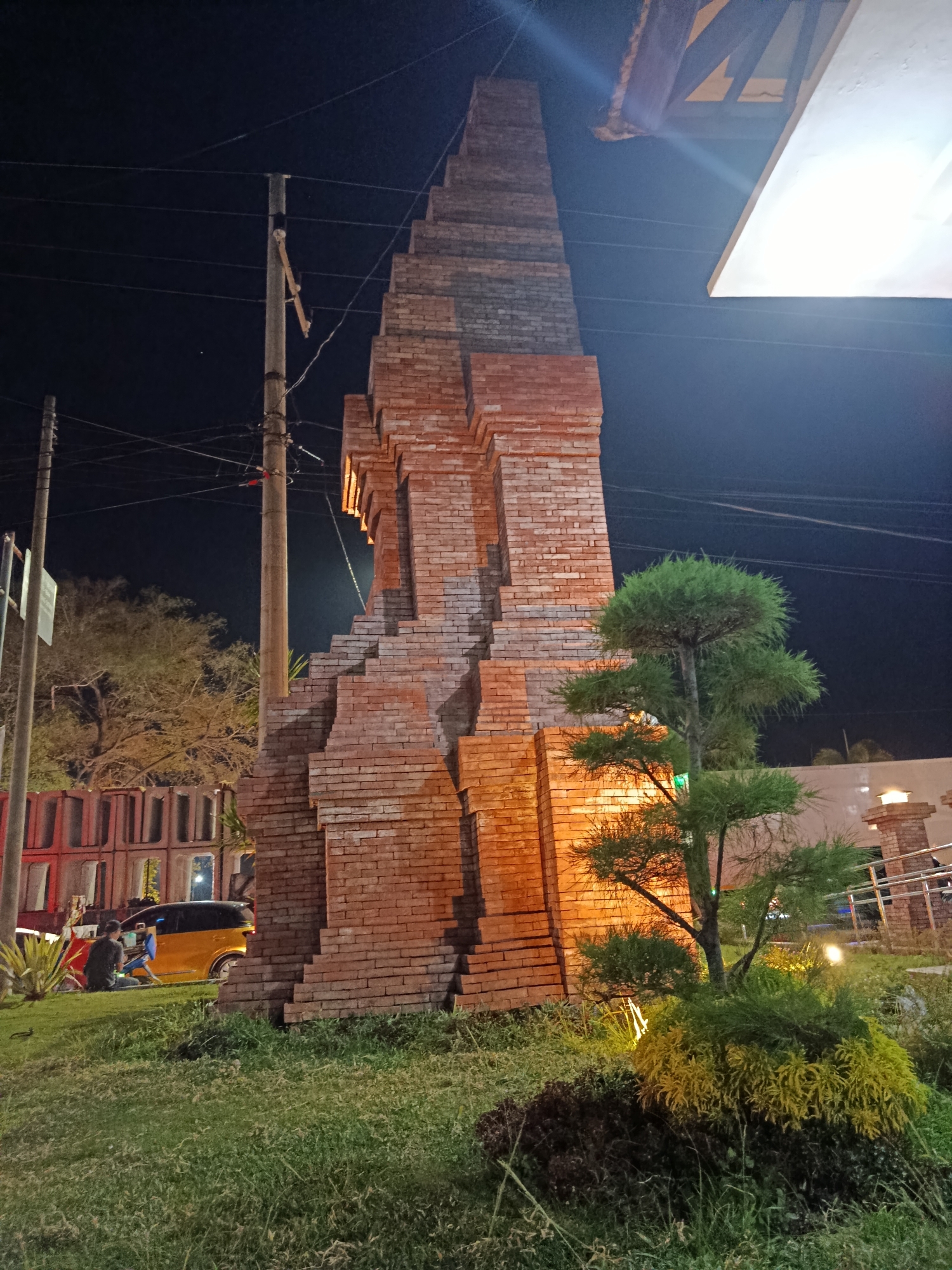 This is a nighttime outdoor photograph taken in Besuki, Indonesia. The main subject is a tall, stepped brick structure, possibly a monument or architectural feature, lit from below, casting a warm glow. In front of the structure, a small, well-maintained garden features a bonsai-like tree and other low-lying, textured plants, with yellow and dark green foliage.
To the left of the brick structure, a utility pole with multiple wires is visible against the dark night sky. Further to the left, a bright yellow car is parked, and a person is seated on the ground near it, possibly interacting with a blue object. Behind them, a row of red and grey rectangular structures with open fronts suggests a building or market stalls.
The scene is illuminated by artificial lights, indicating it is nighttime. The sky is dark and clear, with no visible weather phenomena. The presence of utility poles, a car, and the style of the background structures hint at an urban or semi-urban environment. The overall mood is calm and quiet, with the lighting creating a somewhat dramatic ambiance. No visible text is present in the image.