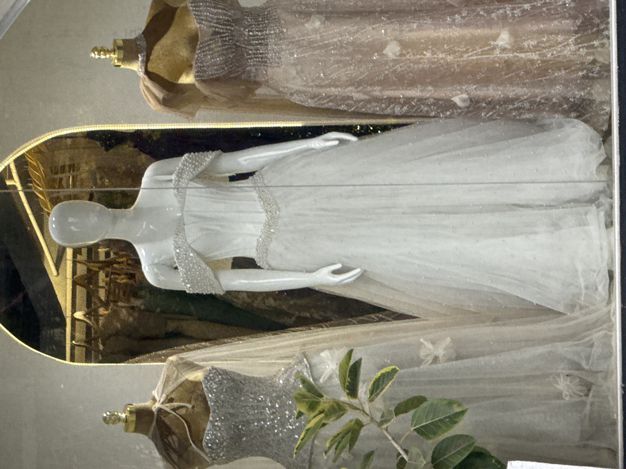 This is a medium-shot, eye-level, full-color photograph taken through a glass window, likely a storefront display. Three mannequins are showcased, each adorned with elaborate gowns. The central mannequin wears a white, strapless ball gown with delicate beading along the neckline and waist, and a flowing tulle skirt. To the left, a mannequin displays a silver sequined gown with a sheer cape. To the right, another mannequin is dressed in a pale pink, floor-length gown with intricate beadwork and a similar sheer overlay. The background appears to be a dimly lit store interior, with more garments visible on hangers. A small plant with green leaves is positioned in the foreground to the left. The overall aesthetic suggests a bridal or formal wear boutique. There is no direct indication of the location being in Davao City, Philippines.