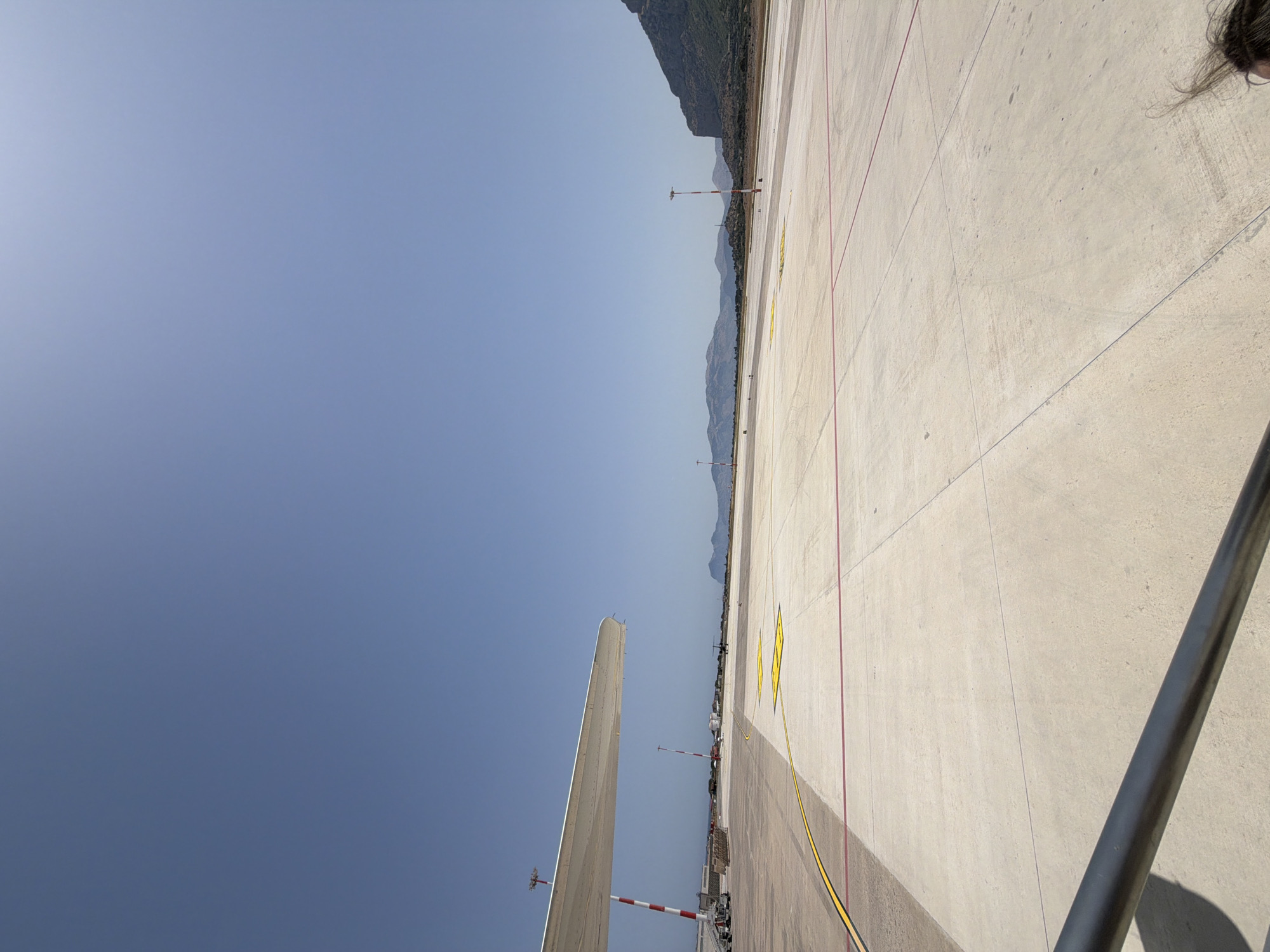 The image depicts an airport tarmac in Cinisi, Italy, under a clear blue sky. In the foreground, a metal railing extends across the bottom of the frame. To the left, a portion of an airplane wing is visible, casting a shadow on the concrete surface. The tarmac is marked with yellow and red lines, and several poles with red and white markings are scattered across the scene. In the distance, mountains rise against the horizon. The image appears to be taken from the perspective of someone on or near an aircraft, perhaps preparing for departure or having just arrived.