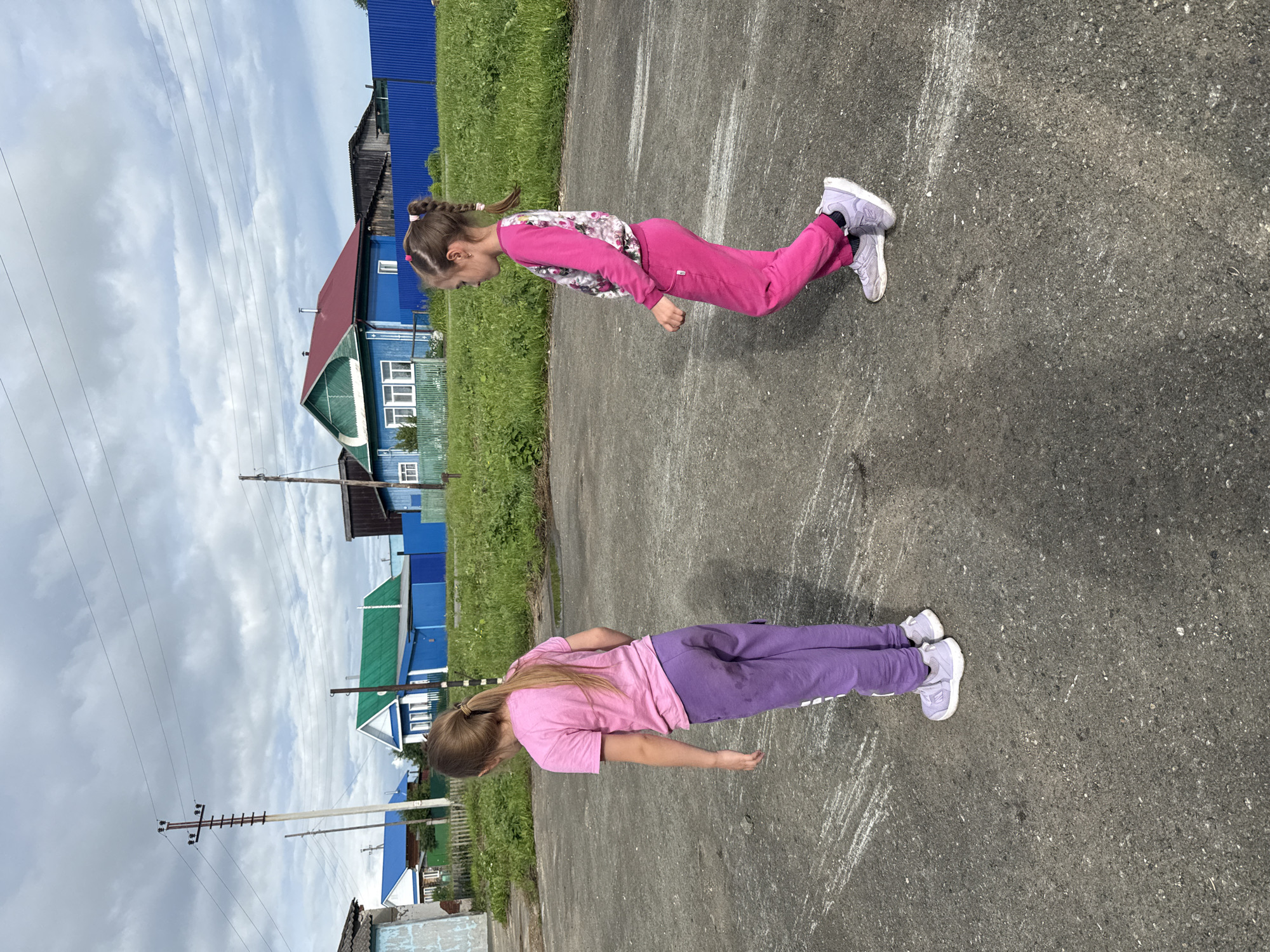 Two young girls are shown performing push-ups on a paved surface, likely a street or playground, in Большой Турыш, Russia. The girl in the foreground wears a pink t-shirt and purple pants, while the girl behind her is dressed in a pink floral long-sleeved shirt and pink pants. Both are wearing light purple sneakers. They are positioned in a push-up stance, with their bodies aligned and arms extended. The ground has white chalk markings.
The background features a grassy area and a row of colorful houses with blue and green roofs, typical of rural Russian architecture. Overhead power lines and utility poles are visible, indicating a residential or village setting. The sky is cloudy, suggesting overcast weather. The scene appears to be outdoors during daylight hours. The girls' focus and the athletic activity suggest a moment of physical exertion and perhaps playful competition. No visible text is present in the image.