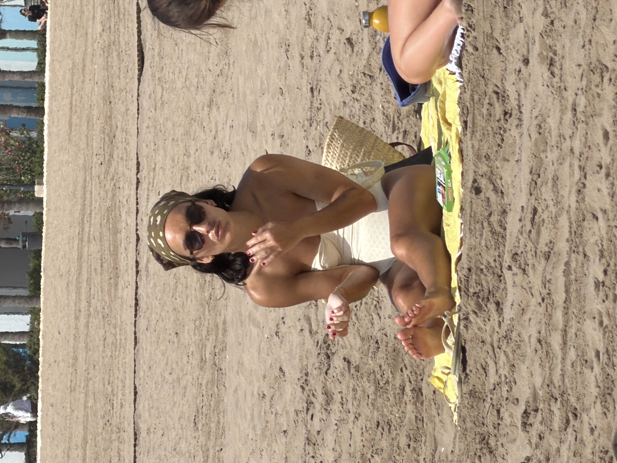 A woman with dark hair tied with a patterned headscarf and sunglasses is sitting on a yellow towel on a sandy beach. She is wearing a white strapless swimsuit and her legs are bent in front of her. Her right hand is raised and open, with fingers spread, as if reaching for something. Her left hand is resting on her lap. To her right, a woven straw bag is visible. Next to the bag, on the towel, is a yellow bottle with a silver cap. In the background, the sand continues, and a blue bin is present, with the legs of another person visible to the right. The environment appears to be a sunny day on a beach with some distant buildings and palm trees.
