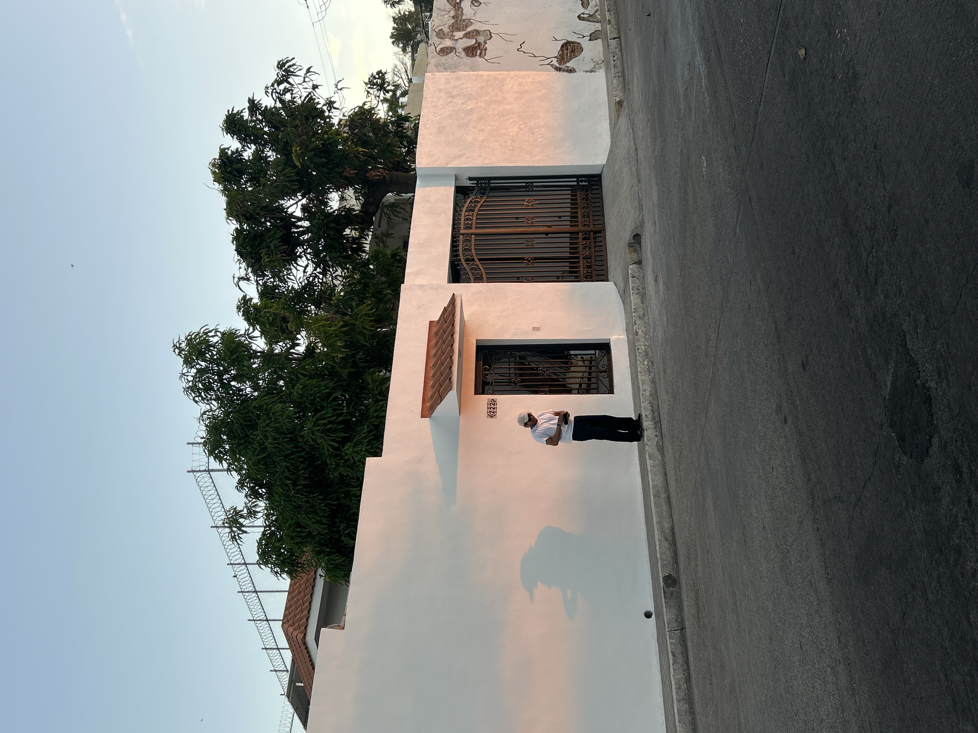 A daytime shot shows a man standing on a sidewalk next to a white wall of a building. He wears a white cap, a white short-sleeve shirt, and dark pants. He appears to be holding something in his hands and looking down at it. His shadow is cast on the wall. The building has a gate and window with decorative metalwork. A number "4222" is visible above the window. Green trees are visible behind the building, along with a fence with barbed wire. The sidewalk borders a paved street. The architecture suggests a location in Mazatlán, Mexico.
