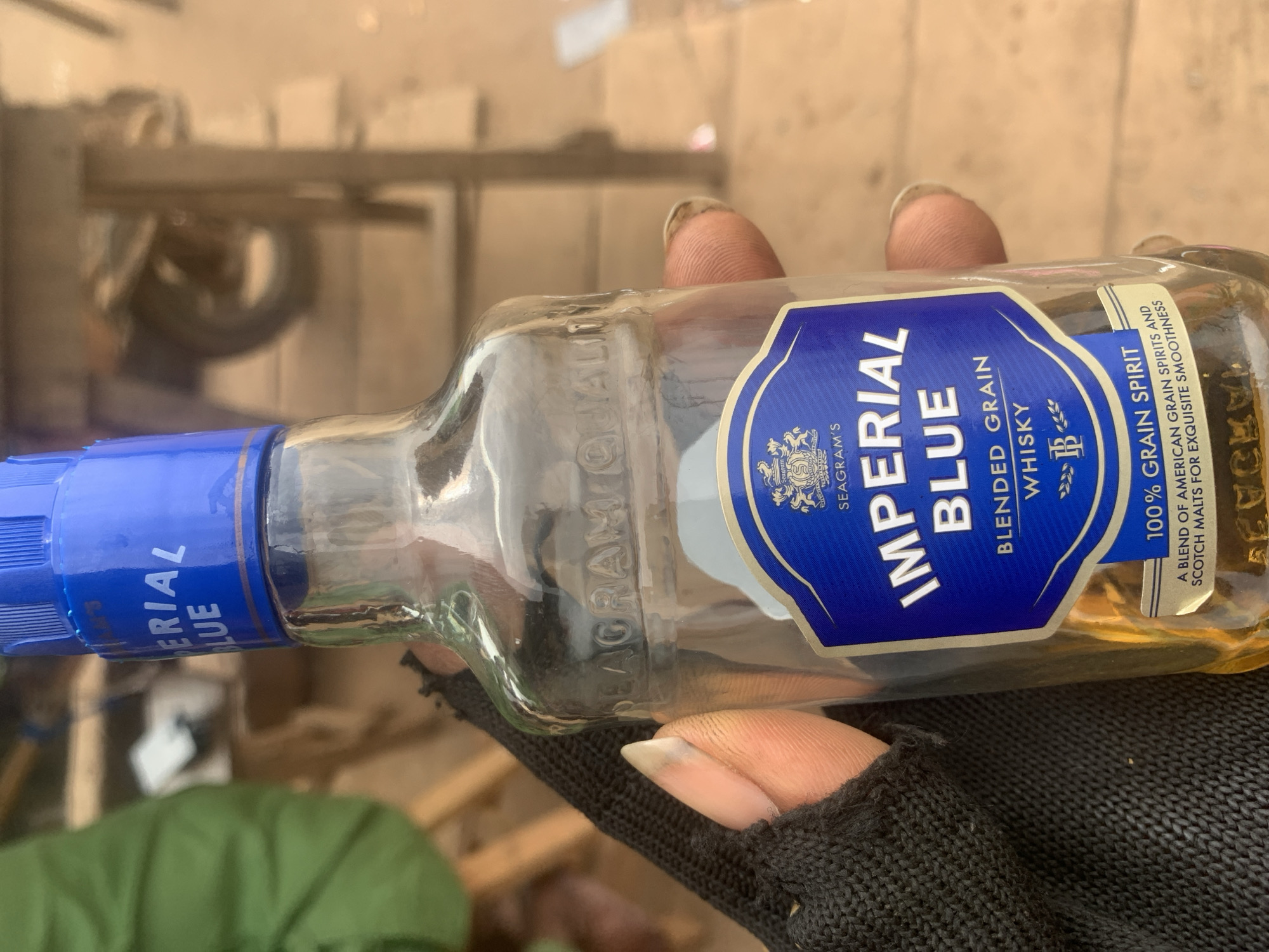 A close-up, eye-level shot shows a hand in a black glove holding a small, clear glass bottle of Imperial Blue blended grain whisky. The bottle has a blue cap and a blue label with the brand name "IMPERIAL BLUE" in large white letters, followed by "BLENDED GRAIN WHISKY". Below this, a silver band reads "100% GRAIN SPIRIT". The background is blurred, but suggests an outdoor environment with wooden structures and possibly a motorcycle. The setting is consistent with the town of Shagamu, Nigeria, known for its marketplaces and informal vendors. The lighting is natural and appears to be daylight. The focus is on the bottle and the hand holding it.