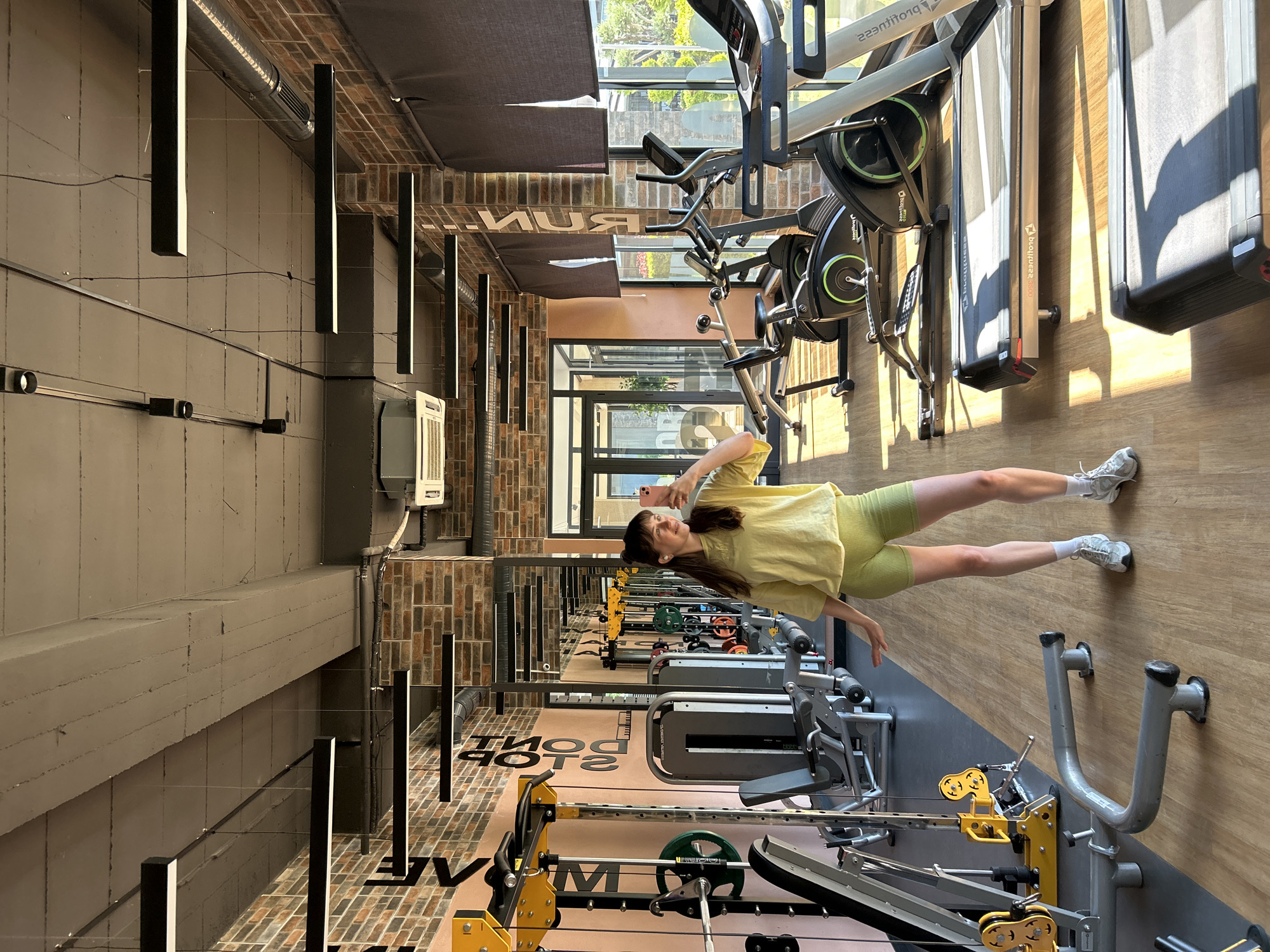 A young woman is taking a selfie with her phone in a gym. She is wearing a light yellow t-shirt and lime green shorts and standing between rows of workout equipment. The gym has weight machines, treadmills and exercise bikes. In the background are windows with sunlight streaming in. The gym floor has the words "TRAIN HARD DON'T STOP." The location is Alanya, Turkey.
