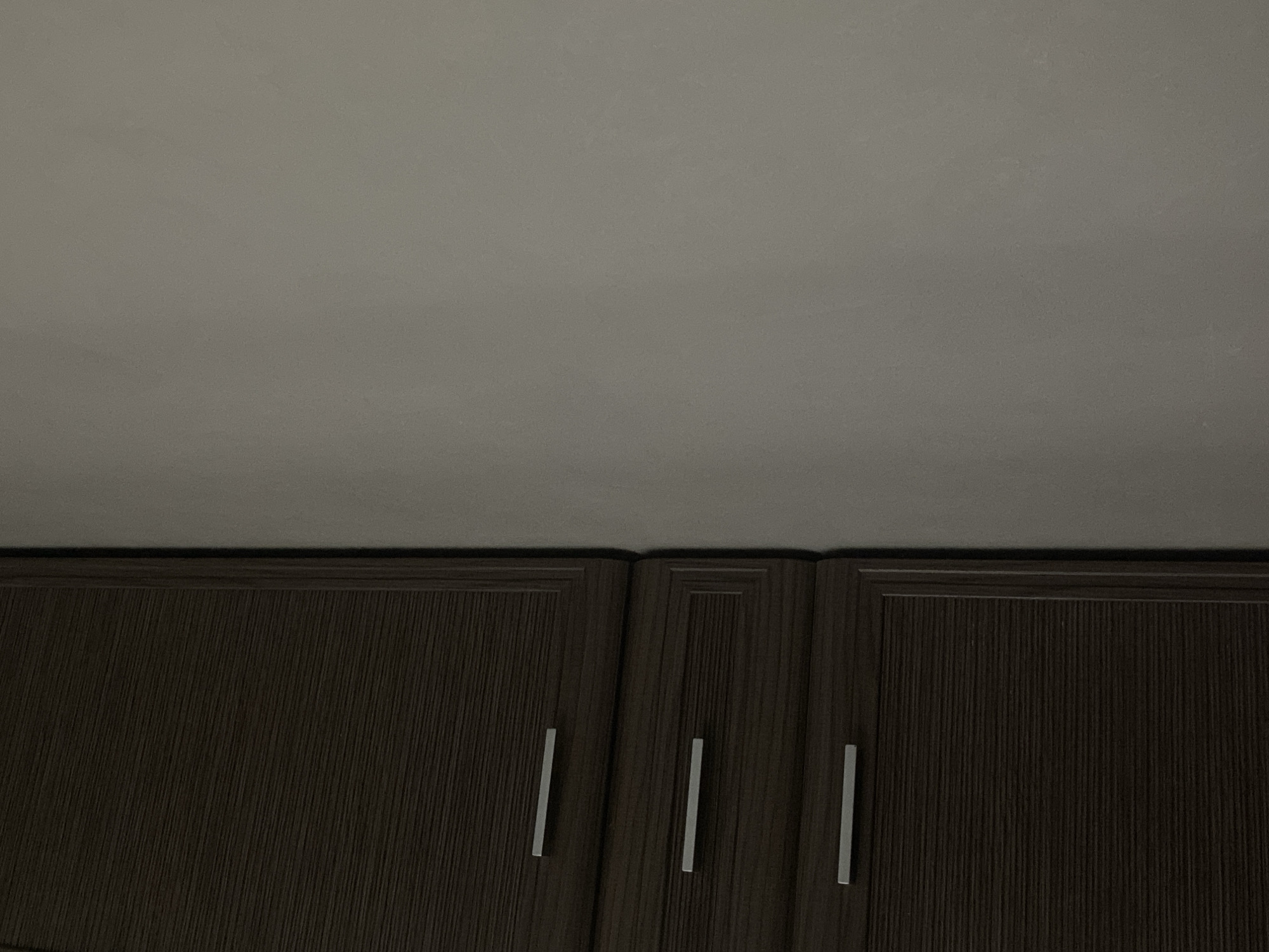 The image shows a close-up view of dark brown wooden cabinets with horizontal wood grain texture. Three silver-colored handles are visible on the fronts of these cabinets, indicating drawers or cabinet doors. The cabinets are positioned on the left side of the frame, meeting a plain, light gray wall on the right. The lighting is dim, casting soft shadows and creating a muted atmosphere. No people, animals, or specific landmarks are present, making the location general and not city-specific. The scene appears to be indoors, likely a room with built-in cabinetry.