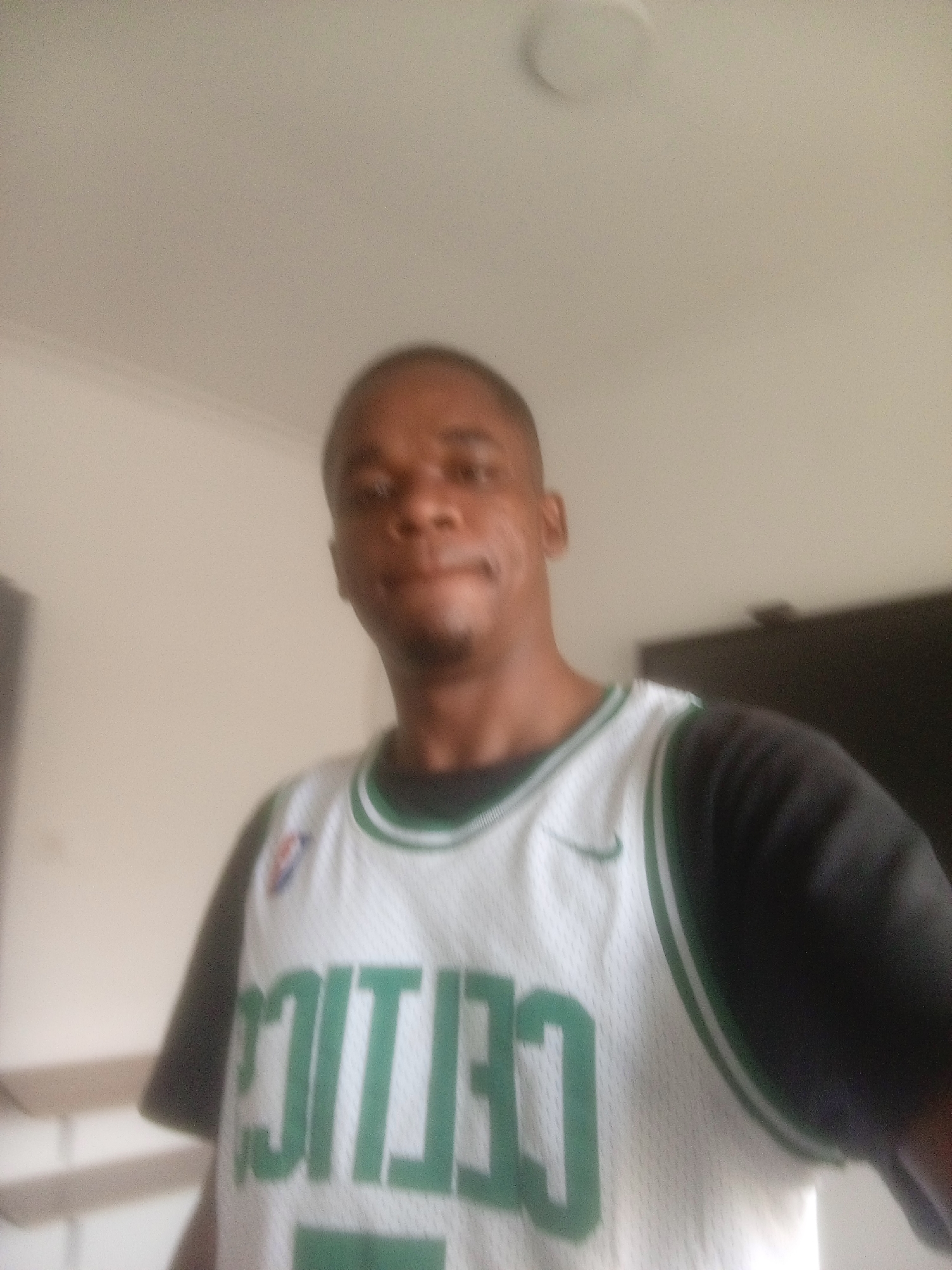 A medium shot of a man wearing a Boston Celtics jersey. He is indoors. The location cannot be verified as being in Abuja, Nigeria.