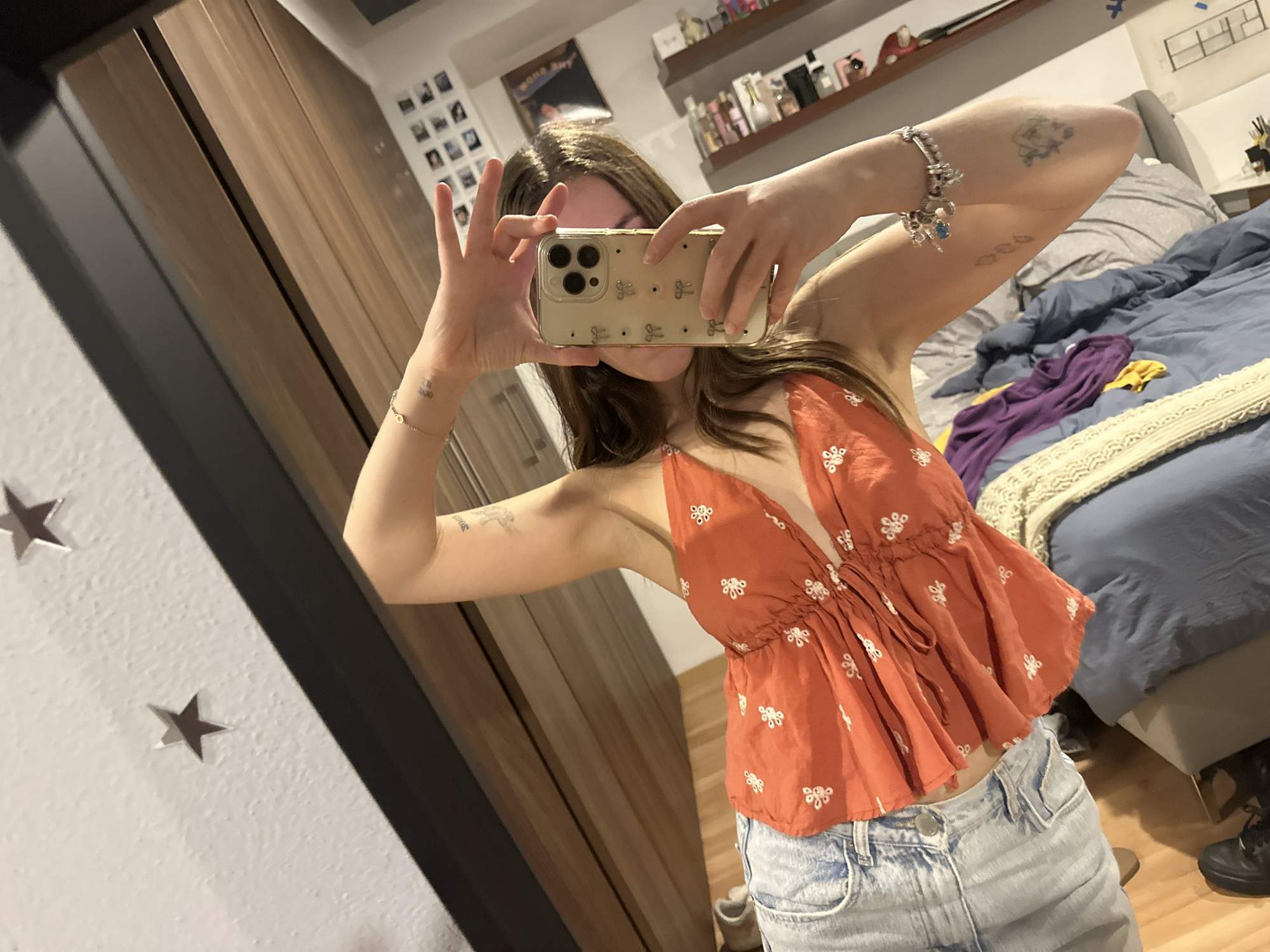 A person is taking a selfie with a beige smartphone with a pattern of bows on its case. The person is wearing a bright orange halter top with small white floral patterns. They are also wearing light-wash denim shorts. The background includes a bed with blue and gray bedding and a light yellow blanket. A wooden wardrobe is visible, and on the wall, there are star decorations. In the background, a shelf is filled with various items, and a collection of small photos are mounted on the wall. The location appears to be a bedroom. The city is not explicitly visible in the image.