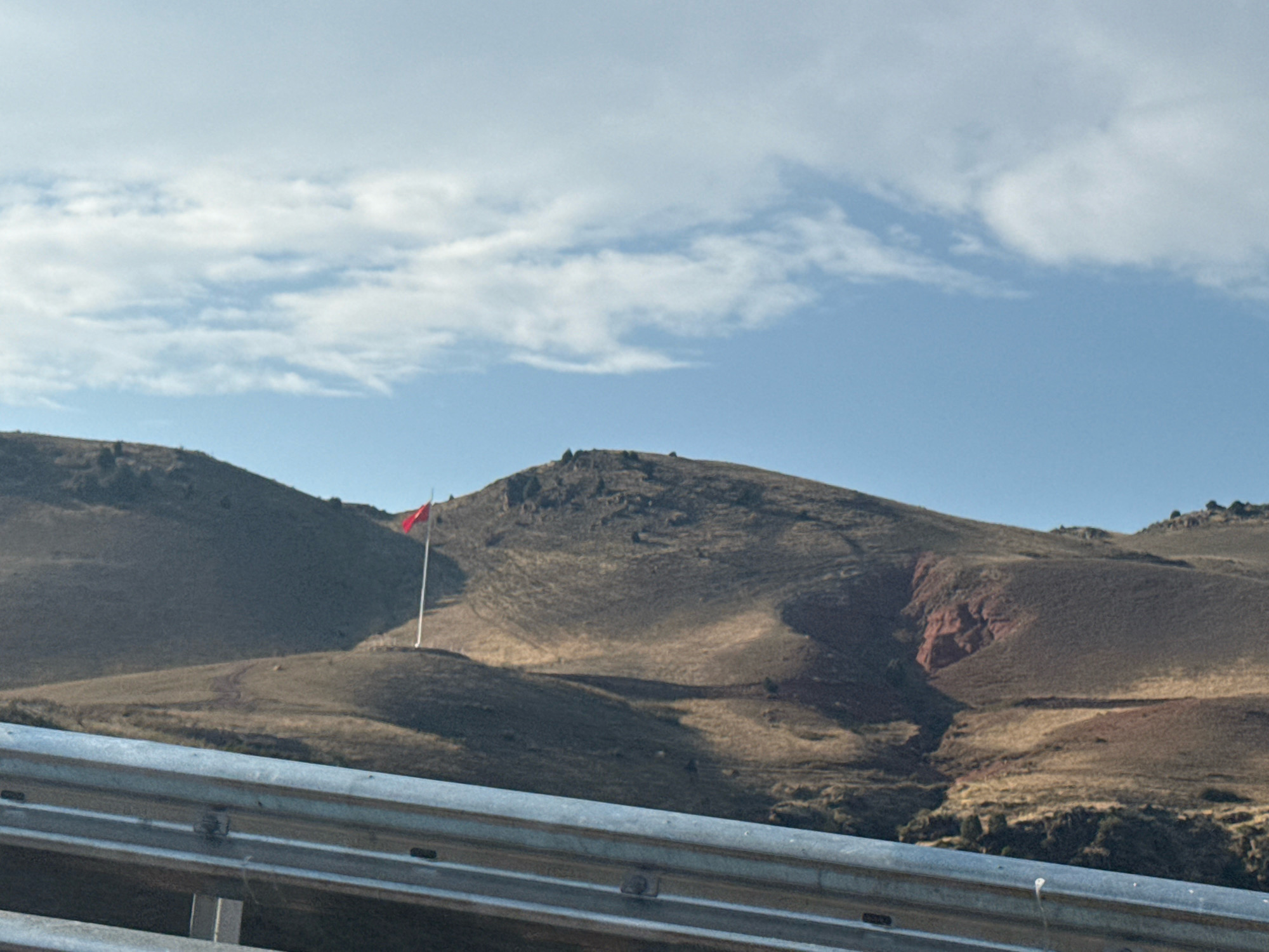 The image depicts a natural landscape with rolling hills under a partly cloudy sky. In the middle ground, a bright red flag is visible on a tall flagpole, positioned on one of the hilltops. The terrain is dry and covered in sparse vegetation. In the foreground, a metallic guardrail runs horizontally across the frame. The scene appears to be a rural or mountainous area.