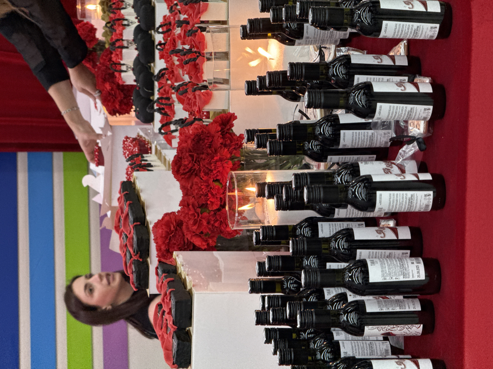 The image shows a scene from what appears to be an event or celebration, with many small wine bottles neatly arranged on a red surface, interspersed with decorative red flowers and candles. The arrangement is on a white table. A woman is visible to the left, near a vertical stripe patterned wall. It is not possible to definitively confirm the location is Yerevan, Armenia, without additional information, although there is an indication on the wine label "TAKAR".