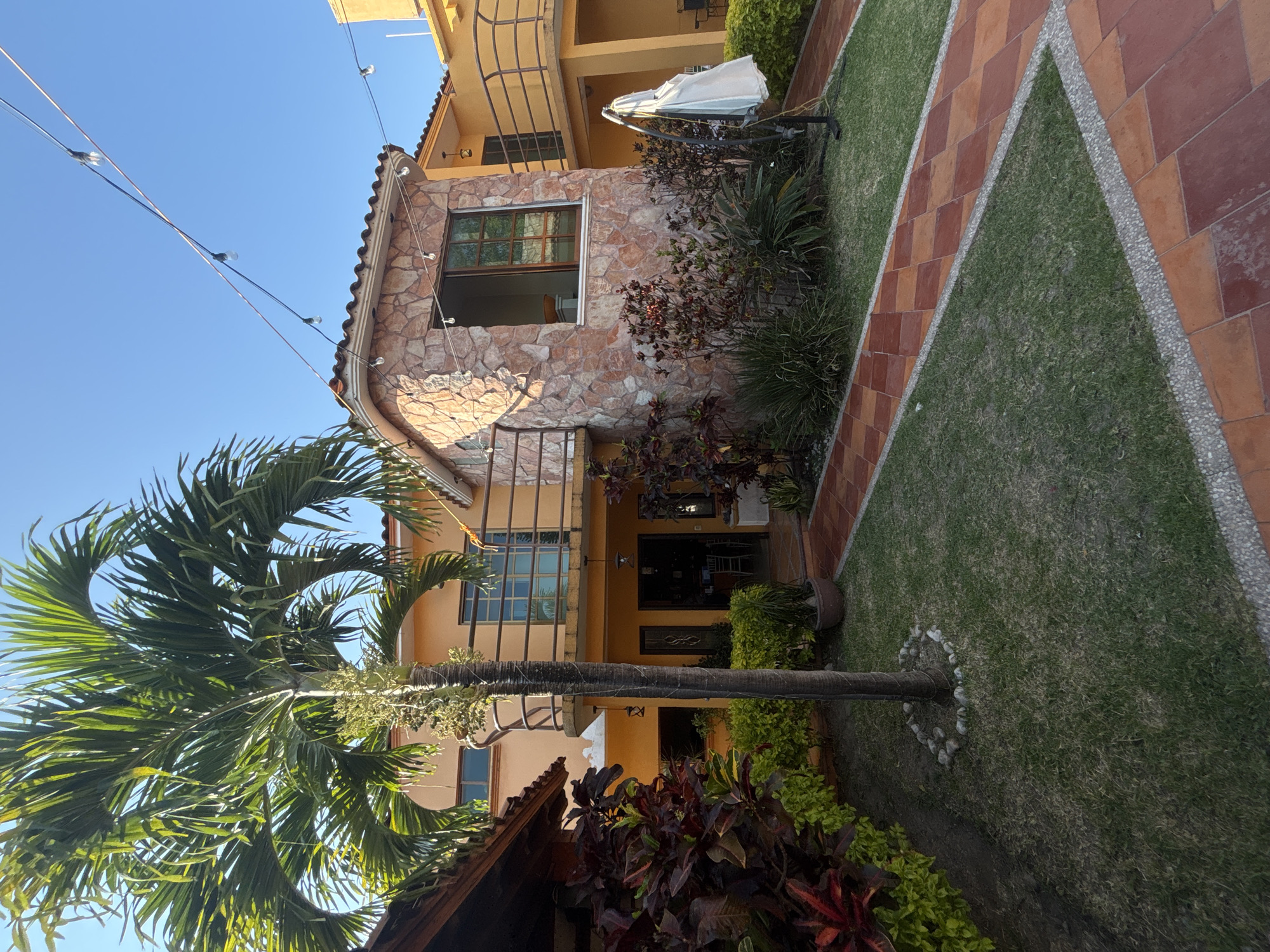 The image shows a two-story house in Chilpancingo, Mexico. The house has a mix of stone and painted stucco facade. A large palm tree is prominently featured in the foreground, partially obscuring the front of the house. The house has a small garden with a tiled walkway and various plants. An outdoor umbrella is visible on the second floor balcony. Overhead electrical wires with decorative lights run across the top left corner of the image.