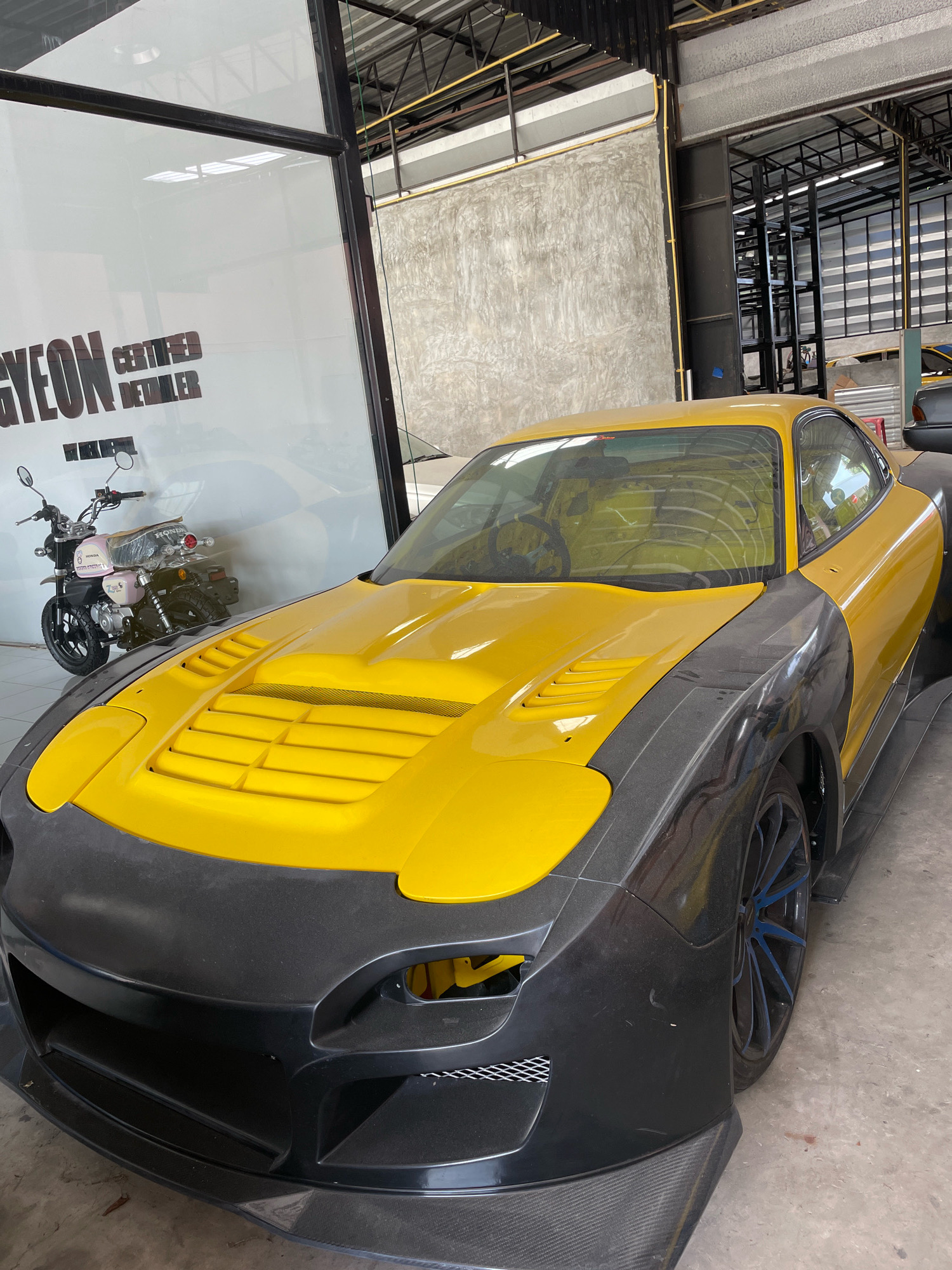 The image shows a bright yellow custom sports car with dark grey fender flares and a black front bumper with visible carbon fiber accents parked inside a garage or workshop in Nong Prue, Thailand. The car has a vented hood. In the background, a pink Honda motorcycle is visible through a glass partition with lettering that reads "GYEON CERTIFIED DETAILER". The environment appears to be an industrial or commercial space with concrete walls and metal shelving structures. The lighting suggests an indoor setting, possibly during daylight hours.