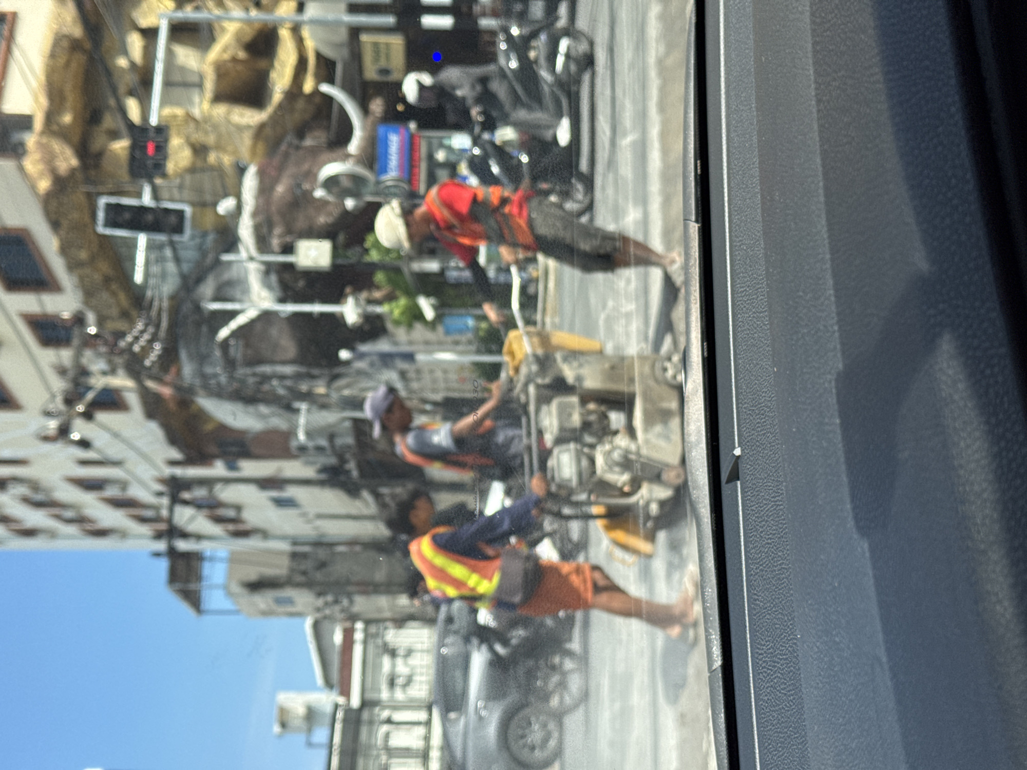 The image shows a street scene in Patong, Thailand, likely on a sunny day, as indicated by the bright sky and shadows. In the foreground, a black car dashboard is visible, suggesting the photo was taken from inside a vehicle.
In the middle ground, construction workers are engaged in road repair. One worker, wearing an orange vest over a blue shirt and orange shorts, operates a concrete saw. Another worker, in a similar orange vest and dark blue shirt, stands nearby. A third worker, wearing a red shirt with an orange vest and a white hard hat, is also present, possibly supervising or assisting. They are working on a paved road, with a piece of construction equipment, a concrete saw, in the center.
In the background, a large building with multiple windows dominates the scene. A prominent, unusually shaped structure, resembling a large, stylized mammoth or rock formation, is situated next to the building, featuring large tusks and a rocky texture. Traffic lights are visible, with a red light illuminated. Several motorcycles and a car are also present on the street, indicating typical urban traffic. The overall impression is one of an active, busy street in a tropical urban environment undergoing construction work.