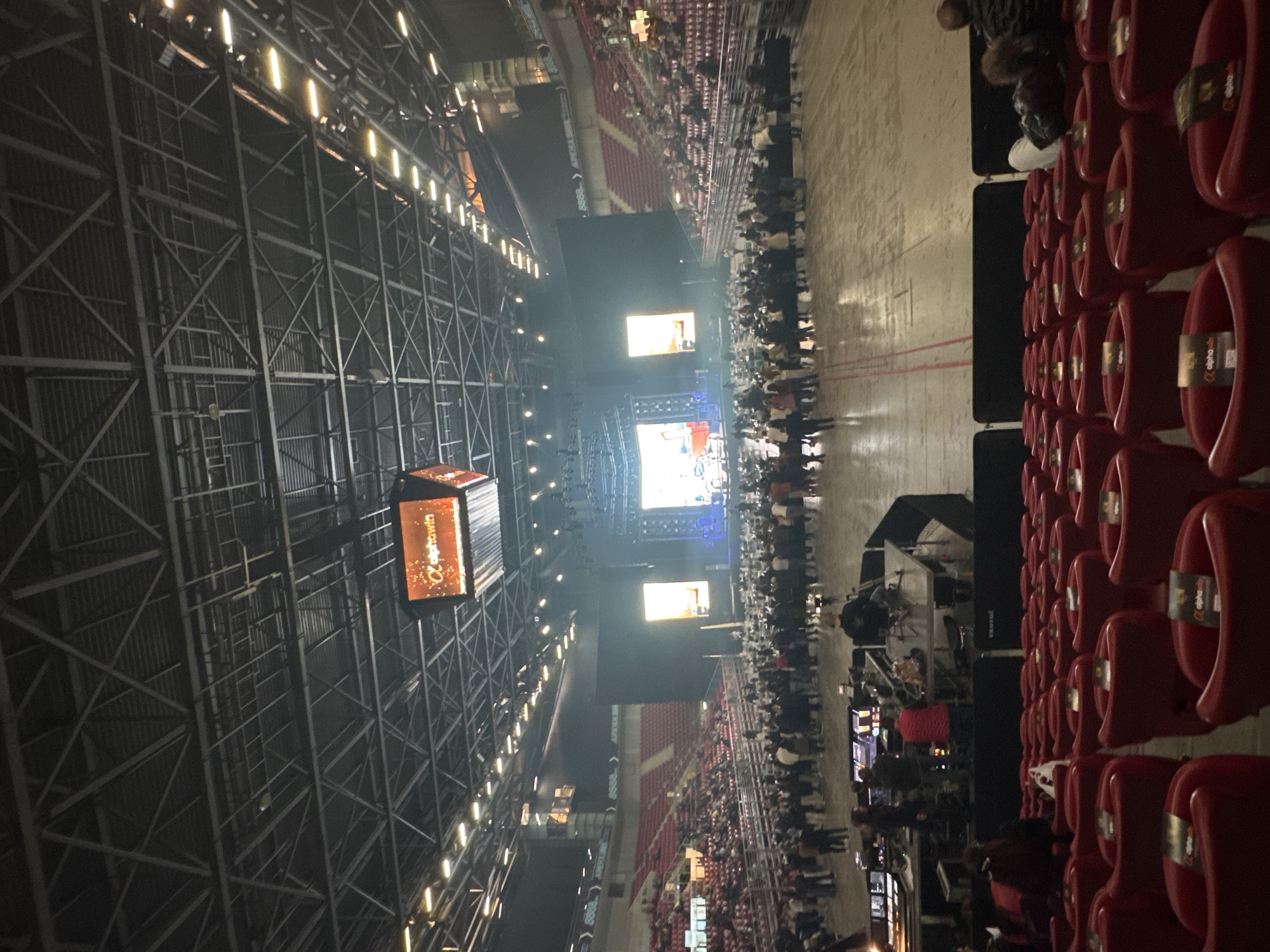 The image captures a high-angle view inside a large indoor arena or stadium. The upper left shows a vast, dark metal truss structure supporting the ceiling, illuminated by rows of bright strip lights.
In the foreground on the right, multiple rows of empty red plastic stadium seats are visible. Many of these seats feature small tags with the "alpha win" logo. Further across the floor level, a large crowd of people is gathered, mostly standing and facing the stage. Additional spectators are seated in the distant red tiered seating.
At the far end of the venue, a brightly lit stage is set up with multiple large video screens displaying colorful visuals. Performers or technical equipment are visible on the stage. A technical or sound booth, equipped with monitors and gear, is positioned on the floor level, closer to the audience.
A cubic display hangs from the ceiling. One visible side of this display shows the text "Unicredit 22," while another side prominently features a golden "alphawin" logo. The venue is dimly lit overall, contrasting with the bright artificial lighting emanating from the stage, screens, and ceiling fixtures. The scene suggests a live event, such as a concert, conference, or performance, taking place indoors.
