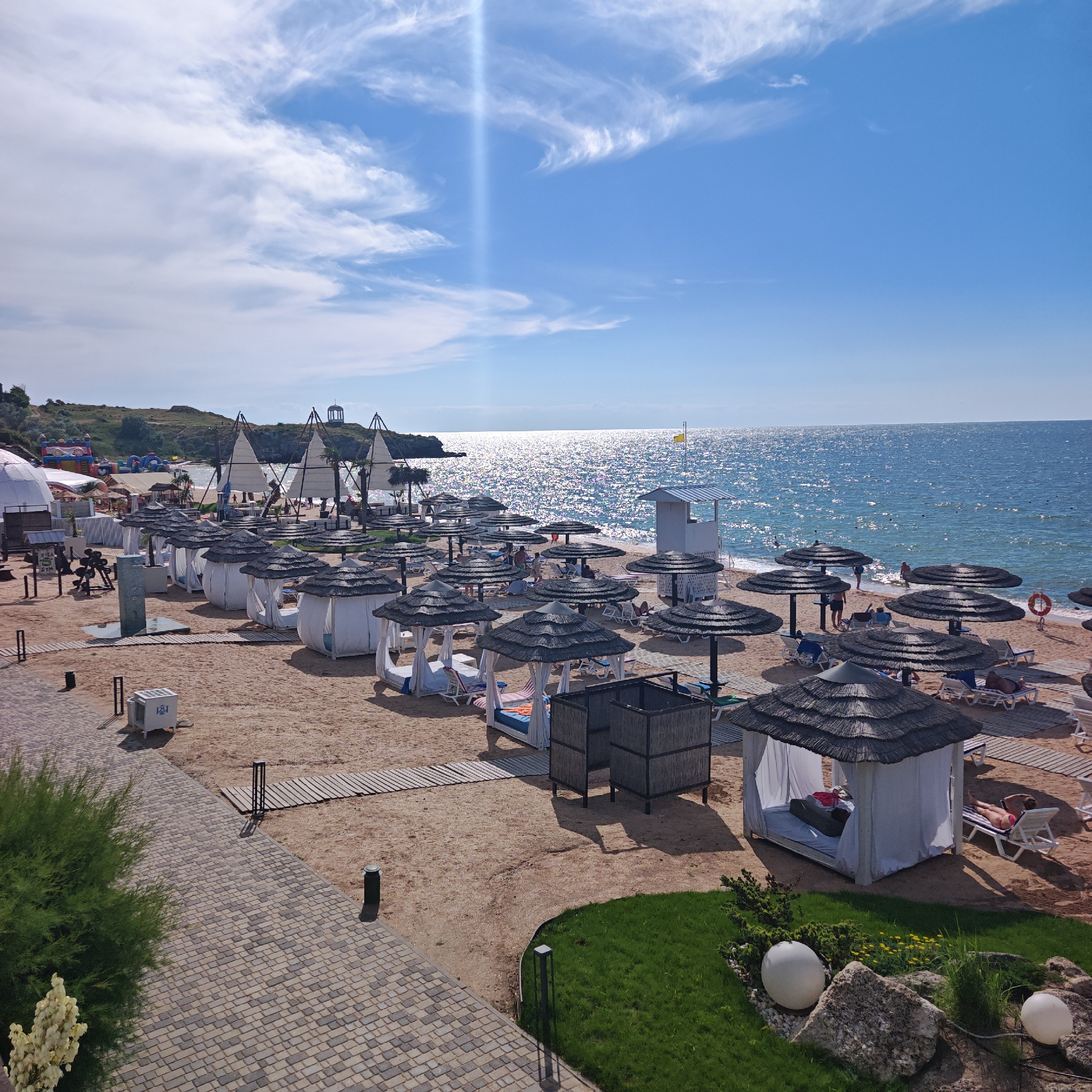This is a sunny, daytime photograph of a resort beach. The beach is lined with numerous thatched-roof cabanas and umbrellas, offering shade to lounge chairs scattered across the sand. In the foreground, a paved walkway and a patch of green grass with decorative spheres and rocks lead to the beach. Further back, several white gazebos with billowing curtains are visible, some occupied by people relaxing.
The sea, shimmering with sunlight, stretches to the horizon under a bright blue sky with scattered white clouds. On the far left of the image, a grassy hillside with some buildings and an inflatable playground can be seen. There's a lifeguard tower and a few people wading in the water. The overall scene conveys a relaxed and inviting atmosphere, characteristic of a seaside vacation destination. No visible text is present in the image.