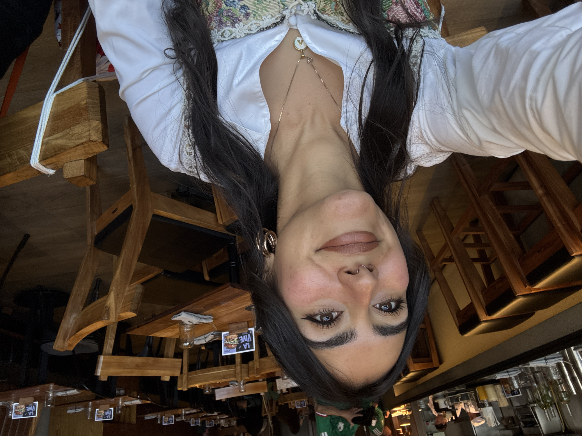 The image shows a woman with long dark hair, hoop earrings, and a gold necklace with a "V" pendant. She is wearing a white blouse with a tapestry-style vest over it. She is indoors, apparently in a restaurant or bar in Mexico City, based on the appearance of wooden tables and chairs and the presence of Mexican soccer jerseys worn by people in the background. There are menus visible on the tables.