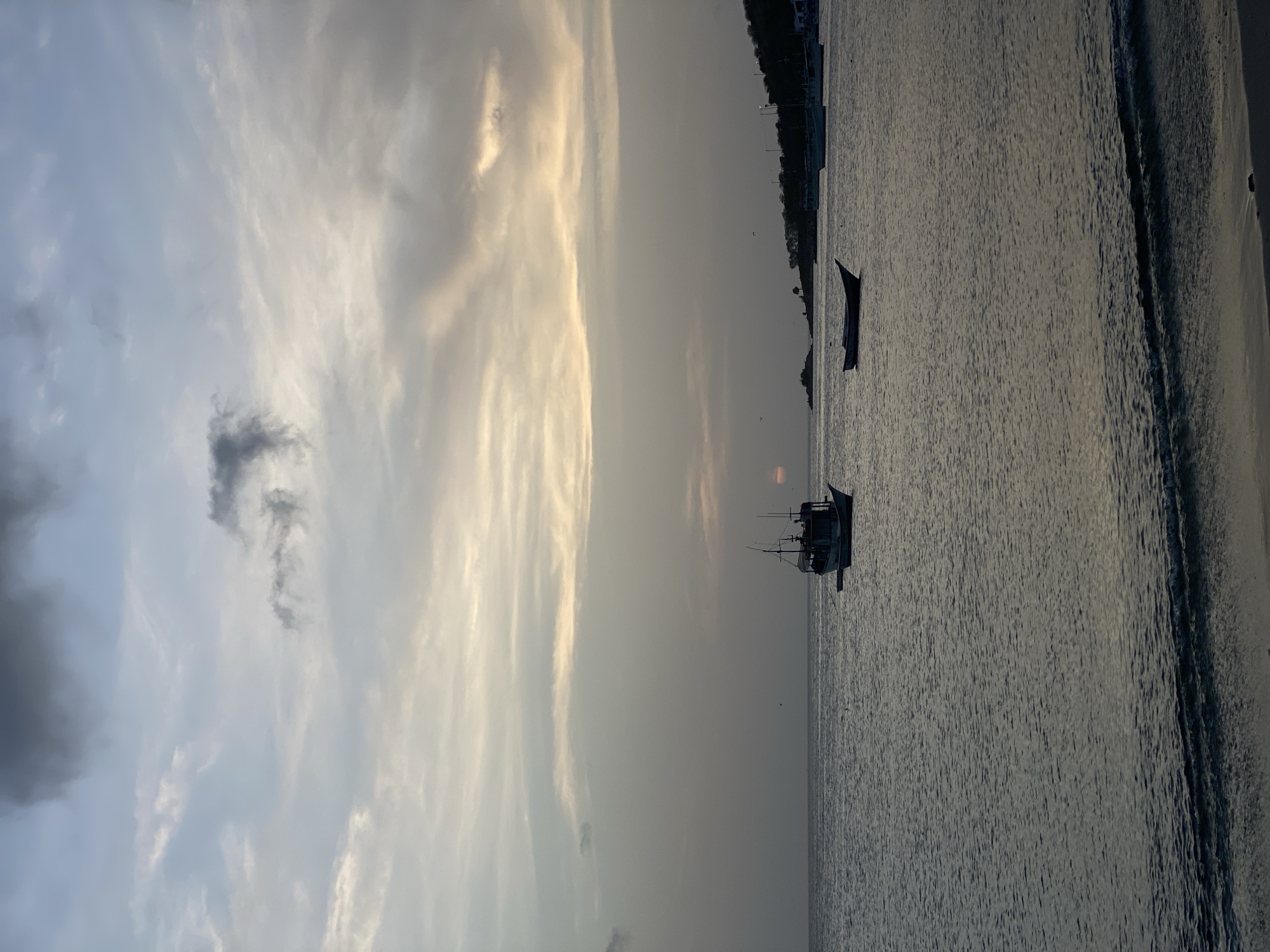 The image depicts a serene coastal scene at sunset in Juan Griego, Venezuela. The sky is a canvas of muted grays and pale yellows, with clouds streaked across the horizon. The sun, a faint orange orb, is partially obscured by the clouds, casting a soft glow. The sea is calm, with gentle waves lapping at a sandy shore in the foreground.
Several boats are visible on the water. In the middle ground, a larger fishing boat with a distinctive silhouette is anchored, appearing to be a focal point of the scene. To its right, a smaller, more slender boat is also at rest on the water. Further in the distance, along the coastline, more boats are lined up, suggesting a harbor or a common mooring area. The coastline itself is lined with trees and buildings, hinting at the presence of a town or village. The overall atmosphere is one of tranquility and natural beauty.