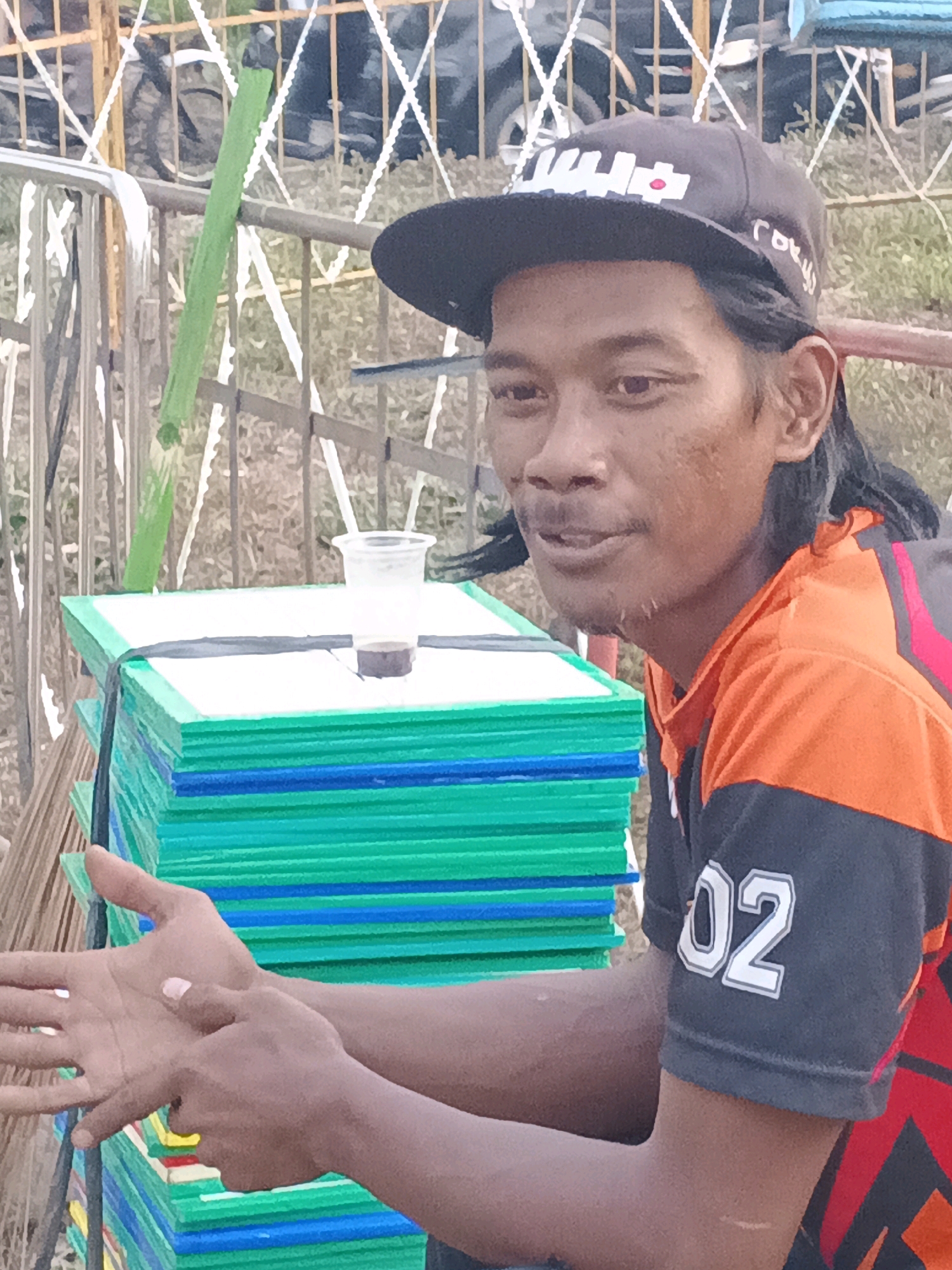 A medium shot captures a young man in Sukabumi, Indonesia, wearing a black baseball cap with white lettering and a red dot, and an orange and black t-shirt with "02" printed on the sleeve. He is sitting or leaning against a stack of colorful rectangular objects, possibly mats, which are green and blue. A clear plastic cup with a dark liquid sits on top of the stack. The man's hands are visible in the foreground, with his left hand open and his right hand touching his left. The background suggests an outdoor setting with a metal fence and some vegetation. The lighting is natural, indicating it is daytime.