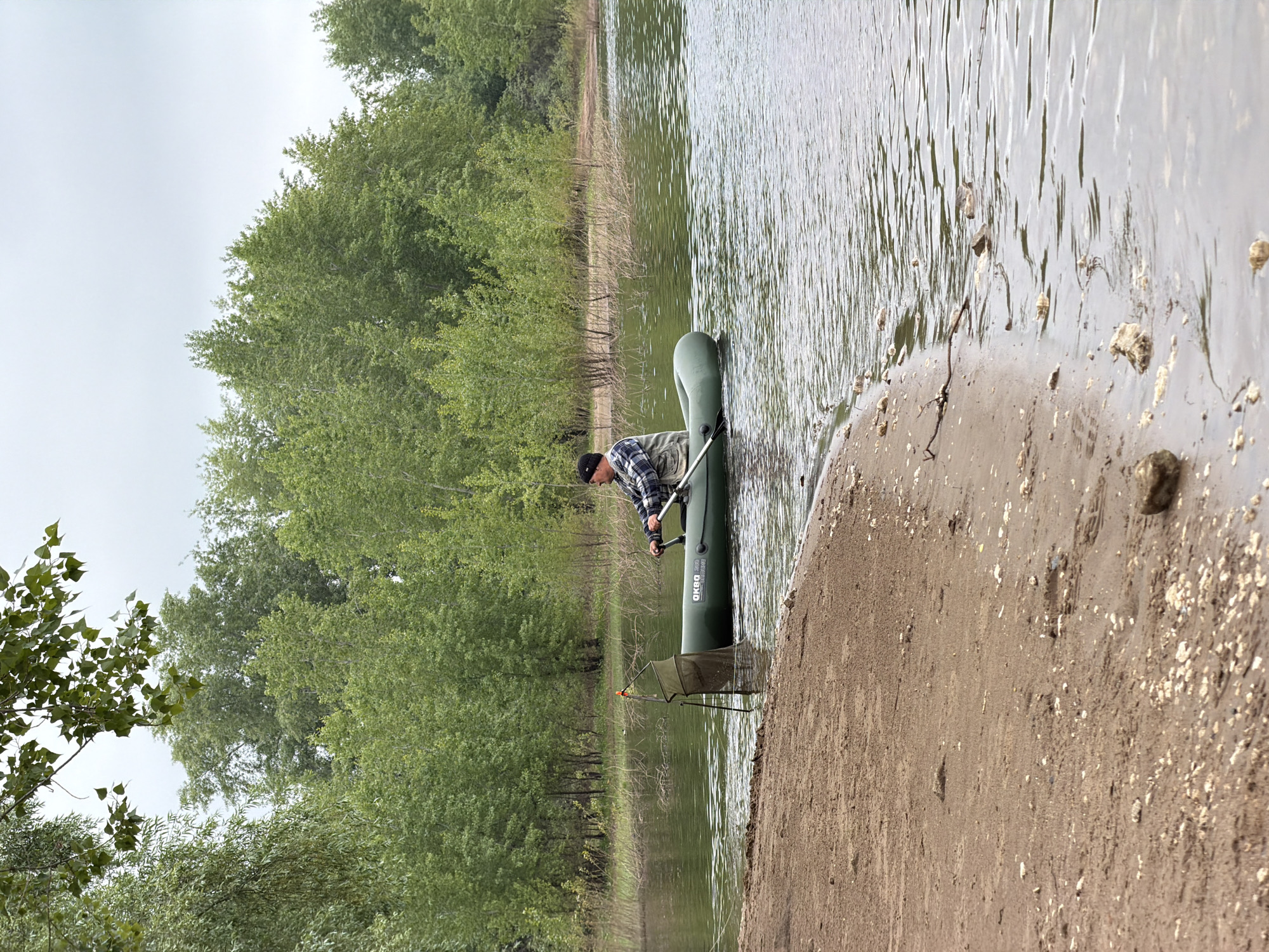 This outdoor photograph captures a man by the edge of a body of water, likely a lake or river, in Chaikovsky, Russia. The scene is set on an overcast day, indicated by the muted light and the expanse of grey sky visible above the treetops.
In the foreground, a sandy and muddy bank slopes gently into the water, scattered with small rocks and debris. A man, partially obscured, is positioned by a green inflatable boat. He wears a dark beanie and a plaid shirt, and is seen holding an oar or pole while interacting with the boat, suggesting he is either preparing to launch it or is getting in. The inflatable boat bears the visible text "АКВА" (AKVA) and "2800" on its side, and is partially submerged in the shallow water. Adjacent to the boat, a fishing keepnet on a stand is set up at the water's edge, implying fishing as the primary activity.
The water surface is covered with subtle ripples, reflecting the overcast sky. The opposite bank and the surrounding landscape are densely populated with vibrant green deciduous trees, creating a natural, verdant backdrop. The overall impression is one of a calm, serene day spent in nature, likely for fishing or boating.