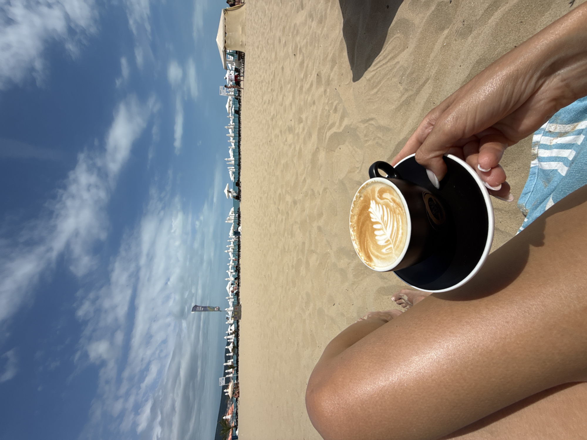 The image depicts a first-person perspective of a person relaxing on a sandy beach in Nesebar, Bulgaria. In the foreground, the person's tanned legs are visible, positioned on a blue and white striped towel. A black cup and saucer with latte art sit in the sand, held by the person's hand. The background shows a crowded beach with rows of white umbrellas and lounge chairs, a blue sky with scattered clouds, and distant mountains. Several people are scattered throughout the scene, engaged in typical beach activities.