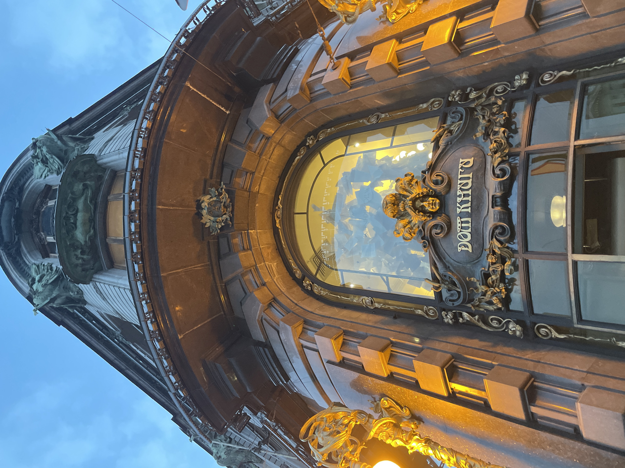 This is a low-angle, close-up shot of the exterior of an ornate building. The building's facade is made of dark, polished stone with elaborate bronze ornamentation. A large, arched window dominates the center of the frame, reflecting the sky and interior lights. The window is framed by detailed bronze scrollwork and a large, sculpted face with a dramatic mane, possibly a lion or mythical creature. Above the window, there are several square, protruding architectural elements.
To the left of the window, the curved facade of the building is visible, adorned with dark, metallic sculptures that appear to be gargoyles or decorative figures. The building's upper edge is lined with a decorative railing. The overall architectural style suggests Art Nouveau or a similar ornate historical style.
Visible text on a dark plaque reads "ДОМ КНИГИ" in Cyrillic script, which translates to "House of Books," indicating the building's likely purpose as a bookstore.
The lighting suggests it is either dusk or dawn, with artificial lights illuminating parts of the building and the sky a muted blue. There is no visible indication of specific weather conditions beyond the sky. The image focuses on the rich details of the architecture, conveying a sense of grandeur and historical significance. No people are visible in the frame.