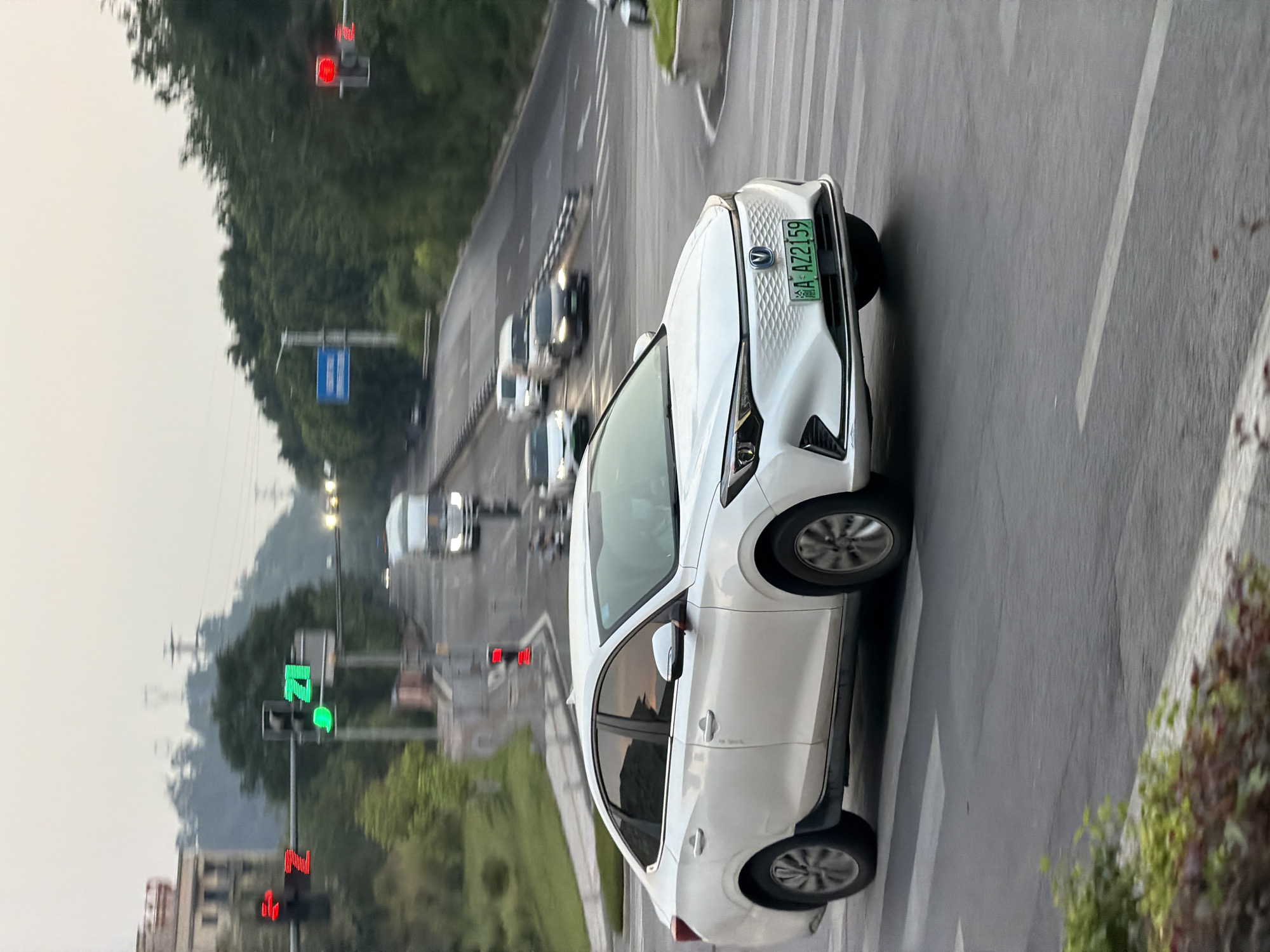 An electric vehicle, identified by a green license plate reading "A AZ2159", is captured in motion on a city street in Jiangjin, China. The vehicle is a white sedan with a distinctive front grille and headlights. The background suggests an urban environment with traffic lights, trees, and other vehicles on the road. The lighting indicates it is either early morning or late afternoon, with overcast skies. The car is positioned in the foreground, traversing a crosswalk. The overall scene depicts a typical urban traffic scenario in Jiangjin.