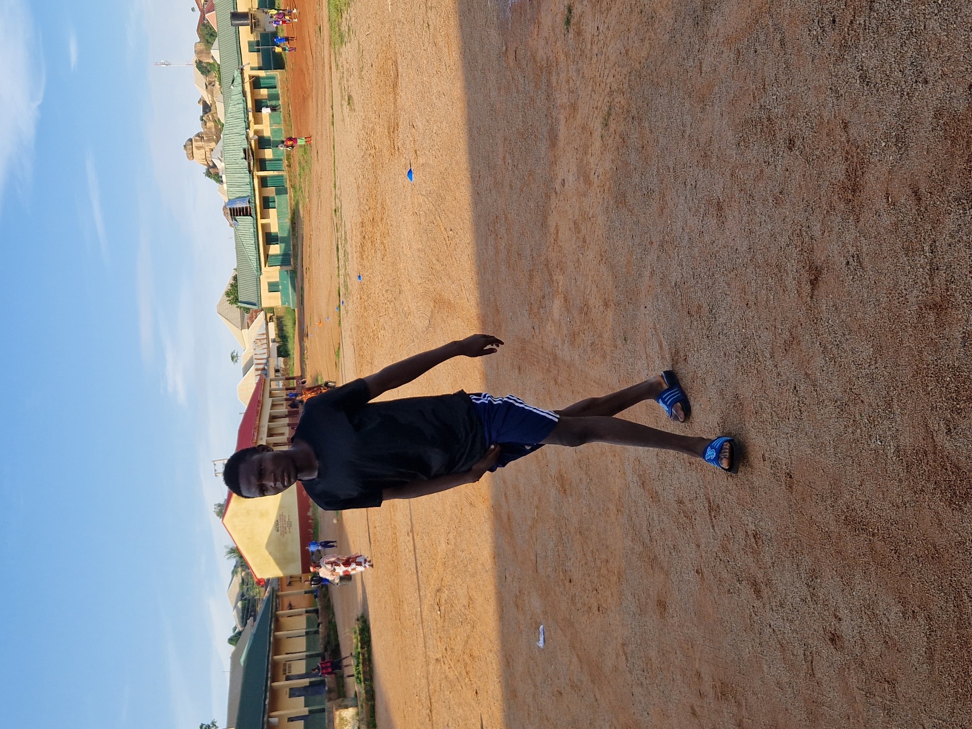 In Kabong, Nigeria, a young Black male is walking across a dirt field in front of a row of light-colored buildings with green trim and red roofs. He is wearing a black t-shirt and blue shorts with white stripes, and blue sandals. In the background, other people are visible, some of whom appear to be playing a game with a ball. The sky is clear and blue. The scene appears to be a school or community area.