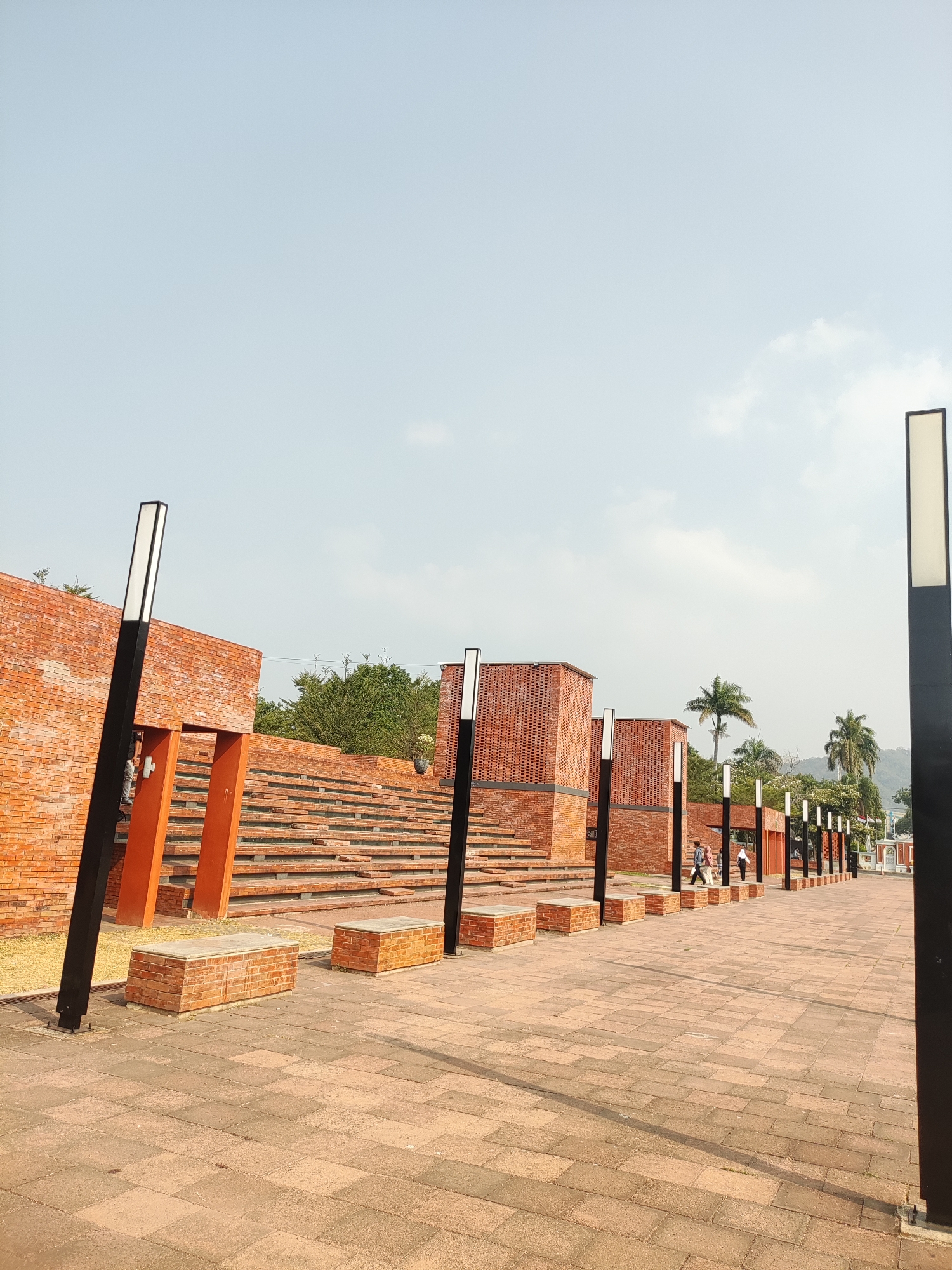 The image depicts an outdoor public space featuring a paved walkway lined with modern black lampposts with rectangular white lights. To the left, there is a large, tiered structure constructed from red bricks, resembling amphitheater seating. Adjacent to the seating area are several low, rectangular benches also made of red brick with stone tops. Further to the left, a section of brick wall forms a facade with an open, orange-framed entryway. The background includes lush green trees and palm trees, with a hazy mountain range visible in the distance. The sky is pale blue with a few scattered clouds. Several individuals are seen walking along the walkway in the middle ground. The scene appears to be the Alun-Alun Majalengka square in Majalengka, Indonesia, a public gathering place.