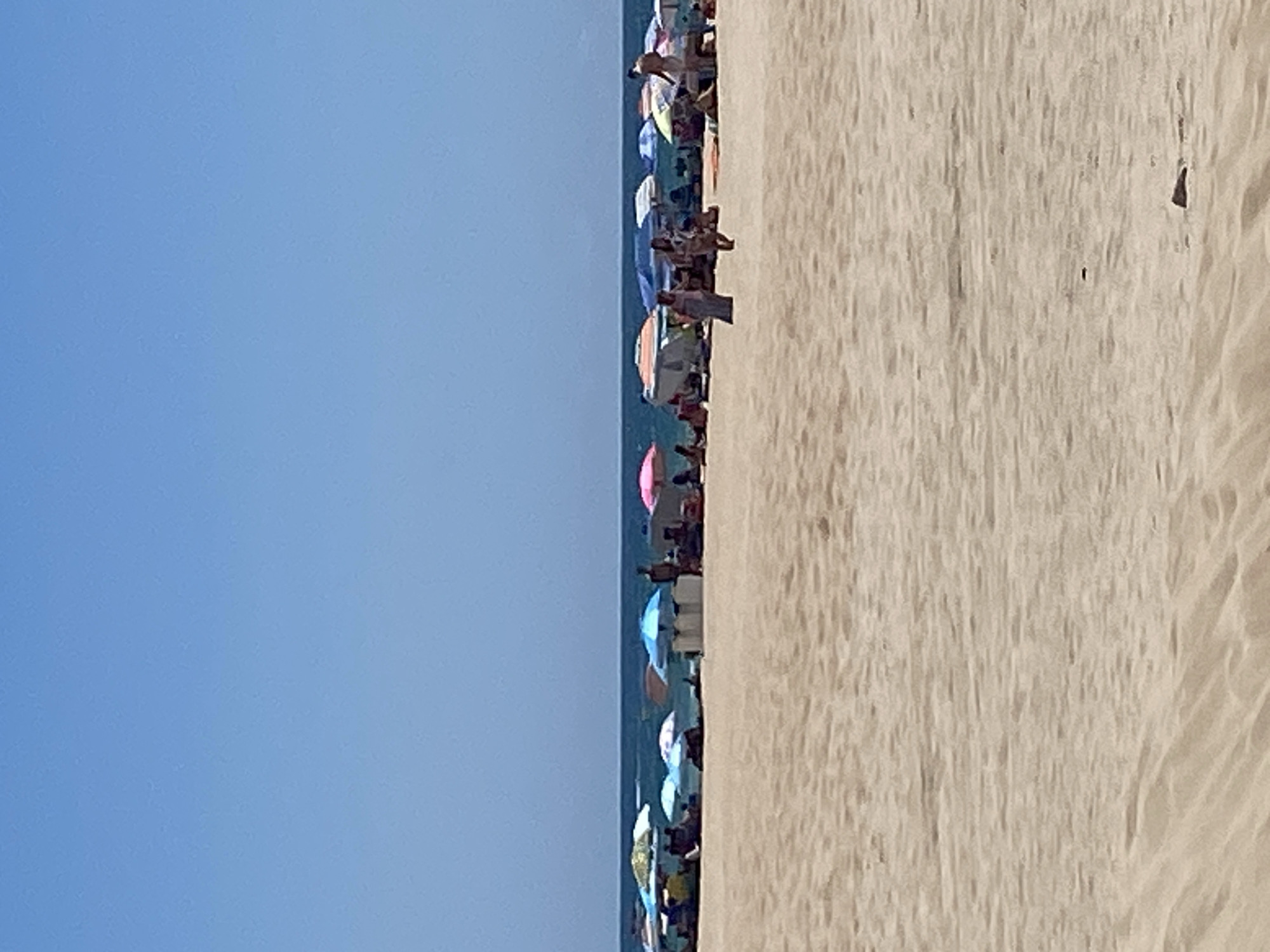 The image captures a sunny day at a beach in Conil De La Frontera, Spain, likely during peak tourist season. The foreground consists of rippled sand, textured by wind and possibly footprints. In the middle ground, a line of beach umbrellas in various colors – including blue, pink, and patterned designs – shades a scattering of people. Some individuals are visible under the umbrellas, while others are standing or walking on the sand. The people appear to be relaxing, socializing, or moving around the beach. In the background, the calm, blue ocean stretches towards the horizon, meeting a clear, bright blue sky. The overall scene conveys a typical summer day at a popular coastal resort.