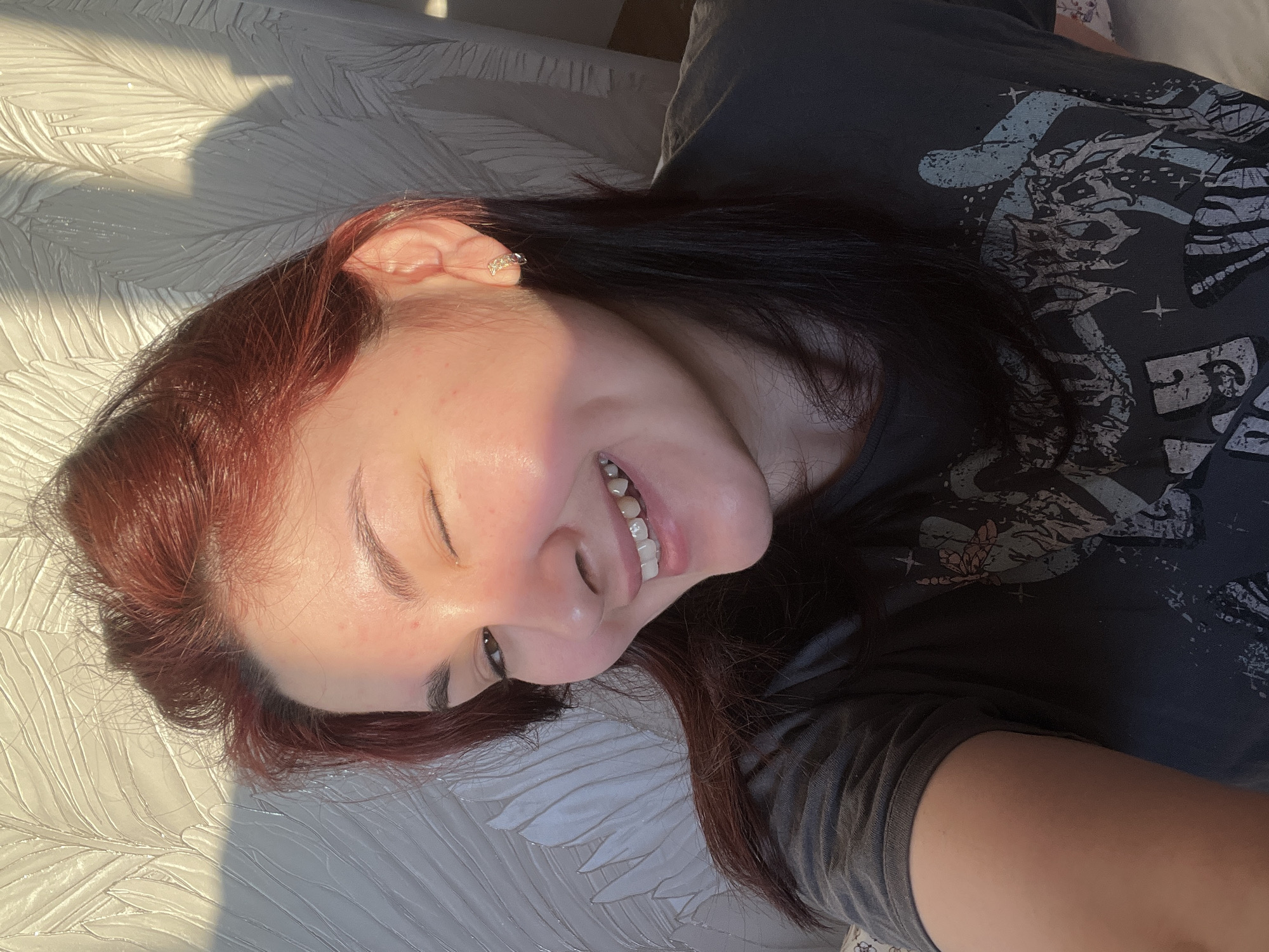 A woman lies on her side on a textured, likely fabric surface, possibly a bed or sofa. She has red hair transitioning to dark near the roots, and is wearing a dark t-shirt with a light-colored graphic design. She is smiling with one eye closed. The lighting suggests a sunny day. It is not possible to determine the location in Neftekamsk, Russia from the image.