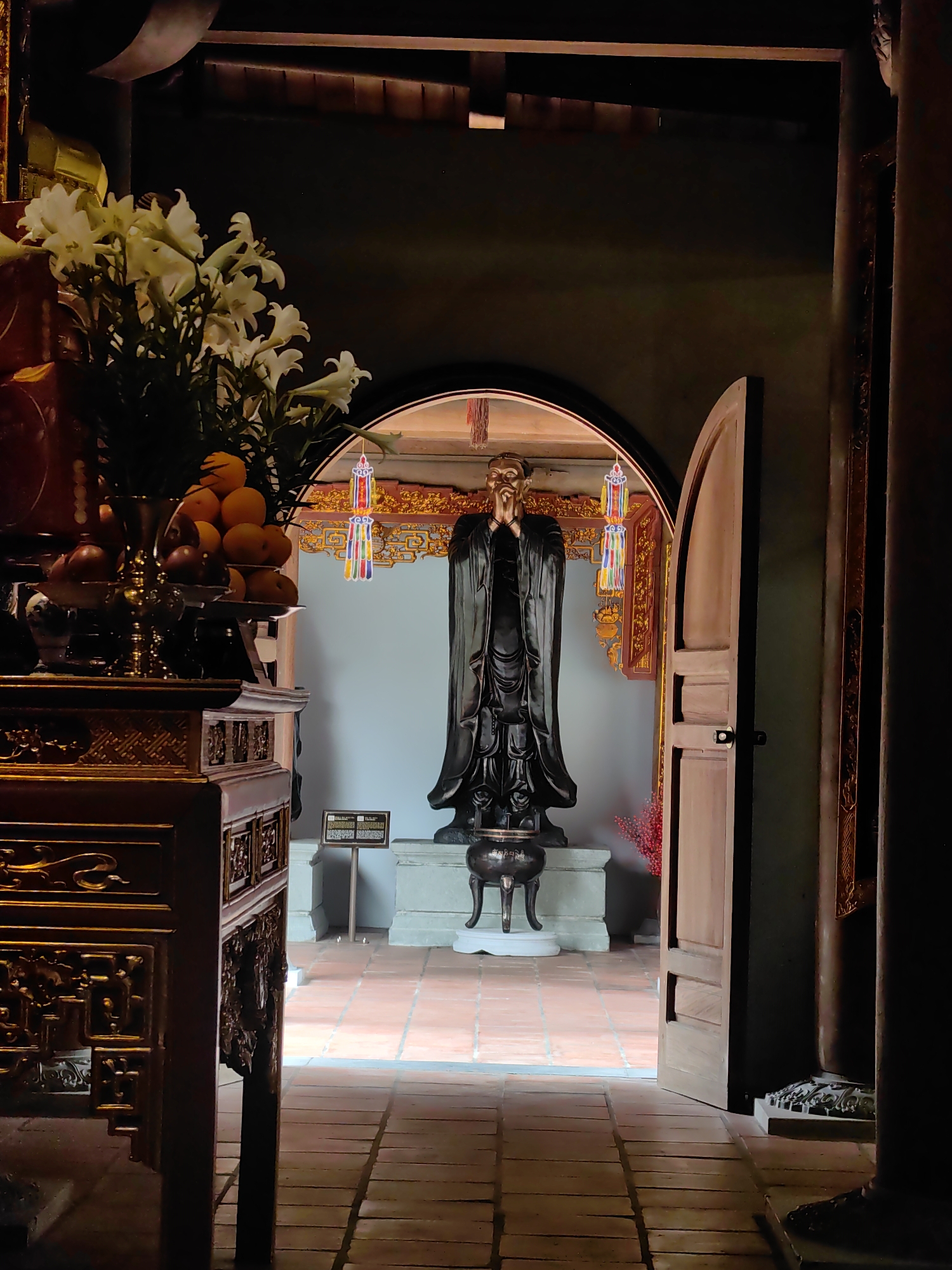 The image captures an interior scene in Hong Gai Ward, Vietnam. A statue of a man stands in a room framed by an arched doorway with an open door, leading from a darker room into a brighter space. The statue, adorned in dark robes and with hands clasped in front of the face, rests on a stand. Decorative elements such as colorful hanging ornaments are visible in the background. In the foreground, there's an ornately carved wooden table with fruits and flowers on top, partially obstructing the view of the doorway and the statue beyond. The floor is tiled in brick-colored squares. The lighting accentuates the statue, while the foreground and surrounding areas are in relative shadow.