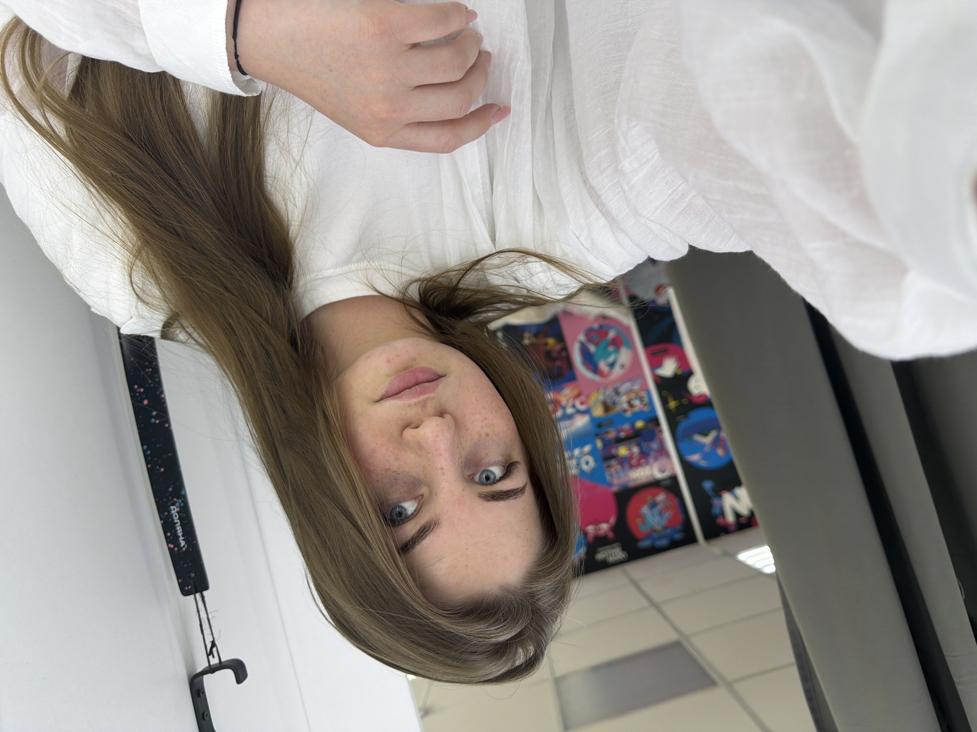 A woman with light skin, blue eyes, and freckles is facing the camera. She is wearing a white button-down shirt and a black bracelet on her wrist. Her light brown hair is styled down. In the background, there is a wall with a series of colorful posters and a ceiling with fluorescent lights. An object with the word "Домаха" and a hook attached is visible on a white wall.
There is no information in the image to support the conclusion that the location is in the city of Chaikovsky, Russia.