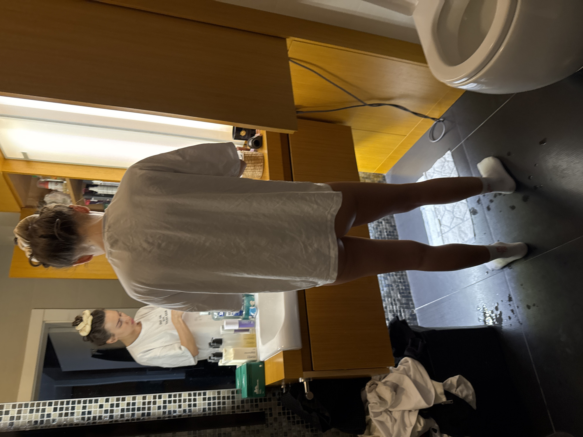 A person is seen from behind, standing in a bathroom in front of a vanity, sink, and mirror in Unknown, Unknown. The individual, appearing to be female, wears an oversized white short-sleeved t-shirt that extends to mid-thigh, revealing bare upper legs, and white ankle socks. Her hair is pulled up into a messy bun, topped with a light-colored, croissant-shaped hair accessory. In the mirror's reflection, the same individual is visible from the front, looking down intently at the countertop as she engages in a personal care routine. The bathroom features a modern wooden vanity with a white rectangular sink. The mirror's left edge is framed by small black and white mosaic tiles. On the vanity to the left of the sink, various toiletries are arranged, including a green box, pump bottles, a purple container, and a clear bottle with blue liquid. To the right of the sink, a woven basket, a black object, and a dark cylindrical container are visible. A tall wooden cabinet with open shelves and closed doors stands to the right of the mirror, displaying various bottles and spray cans. The floor is covered with dark grey or black tiles, with some lighter patterned tiles near a partially visible white toilet on the far right. Several puddles and drops of water are scattered on the dark tiled floor, particularly between the person's feet and a dark laundry basket filled with white clothing situated to the left. Dark electrical cables run along the floor from behind the right side of the vanity.