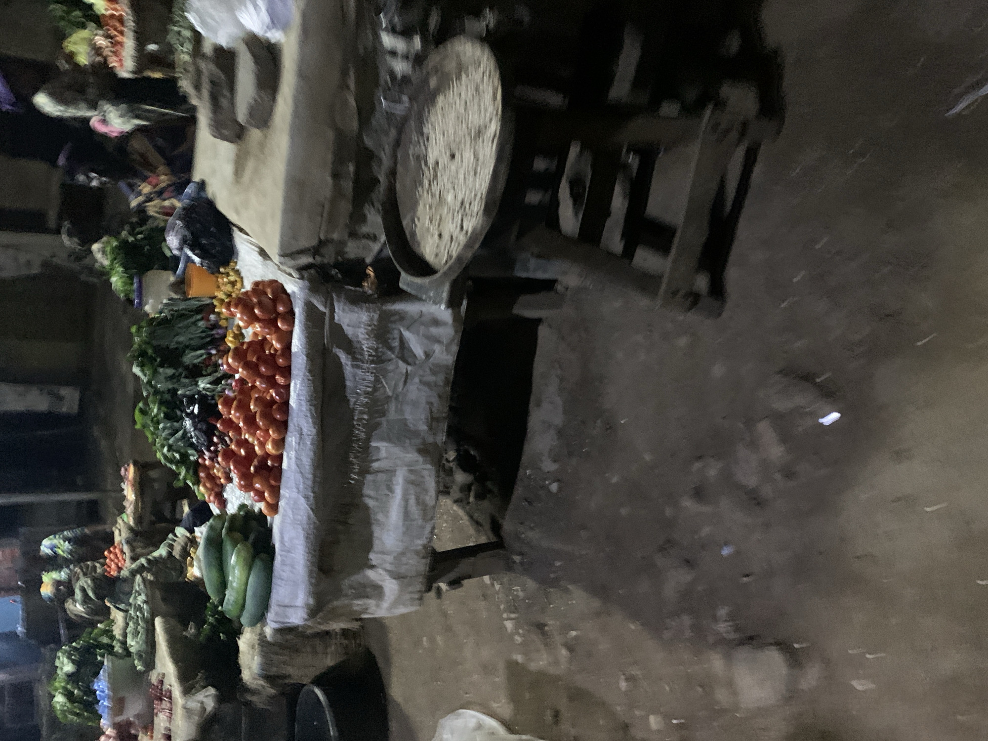 The image depicts a produce stand in what appears to be an outdoor market in Rafin Par, Nigeria. The stand is laden with fresh vegetables, including tomatoes, cucumbers, and leafy greens. A scale rests on the table next to the vegetables. A pigeon is perched near the produce. The environment is dimly lit, suggesting it is either early morning or late evening. The ground is uneven and possibly dirty. Other stands or structures are visible in the background.