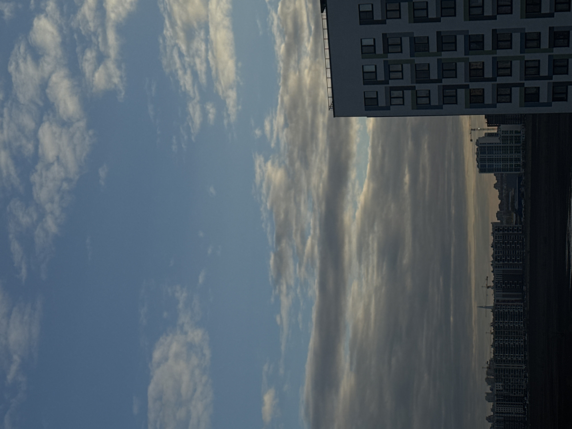 The image presents a broad urban landscape of Saint Petersburg, Russia, under a dramatic sky. The sky occupies the majority of the frame, showcasing varied cloud patterns. On the left side, bright white, fluffy clouds are scattered across a clear blue expanse. Towards the right, the clouds transition into more elongated, streaky formations with darker, grey undertones, suggesting depth and movement.
In the mid-ground, on the far right, a portion of a modern, multi-story building is prominently featured. It exhibits a distinctive architectural style with a repetitive geometric pattern outlining its numerous windows, primarily in shades of grey and white. Below this structure, a distant city skyline stretches across the horizon. It comprises numerous high-rise buildings, many of which appear to be under construction, evidenced by visible cranes. The bottom of the image reveals a dark, horizontal strip, likely a body of water or a distant landmass, extending up to the city's base.
The lighting conditions suggest either early morning or late afternoon, with soft, ambient light illuminating the clouds and accentuating the outlines of the urban structures. The overall atmosphere is expansive and serene. No discernible text is visible within the image.