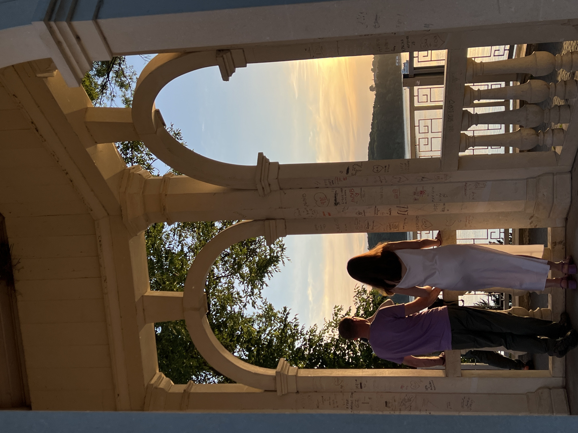 The image shows a couple standing in a white gazebo, holding hands and looking out at a sunset over a body of water. The gazebo has arched openings and is decorated with graffiti. The couple is seen from behind. The man is wearing a purple t-shirt and dark pants, while the woman is wearing a white slip dress. In the background, there is a treeline and a boat on the water. The sun is setting, casting a warm glow over the scene. This is likely the "Love Bay" gazebo in Ternopil, Ukraine, a popular spot for couples.
