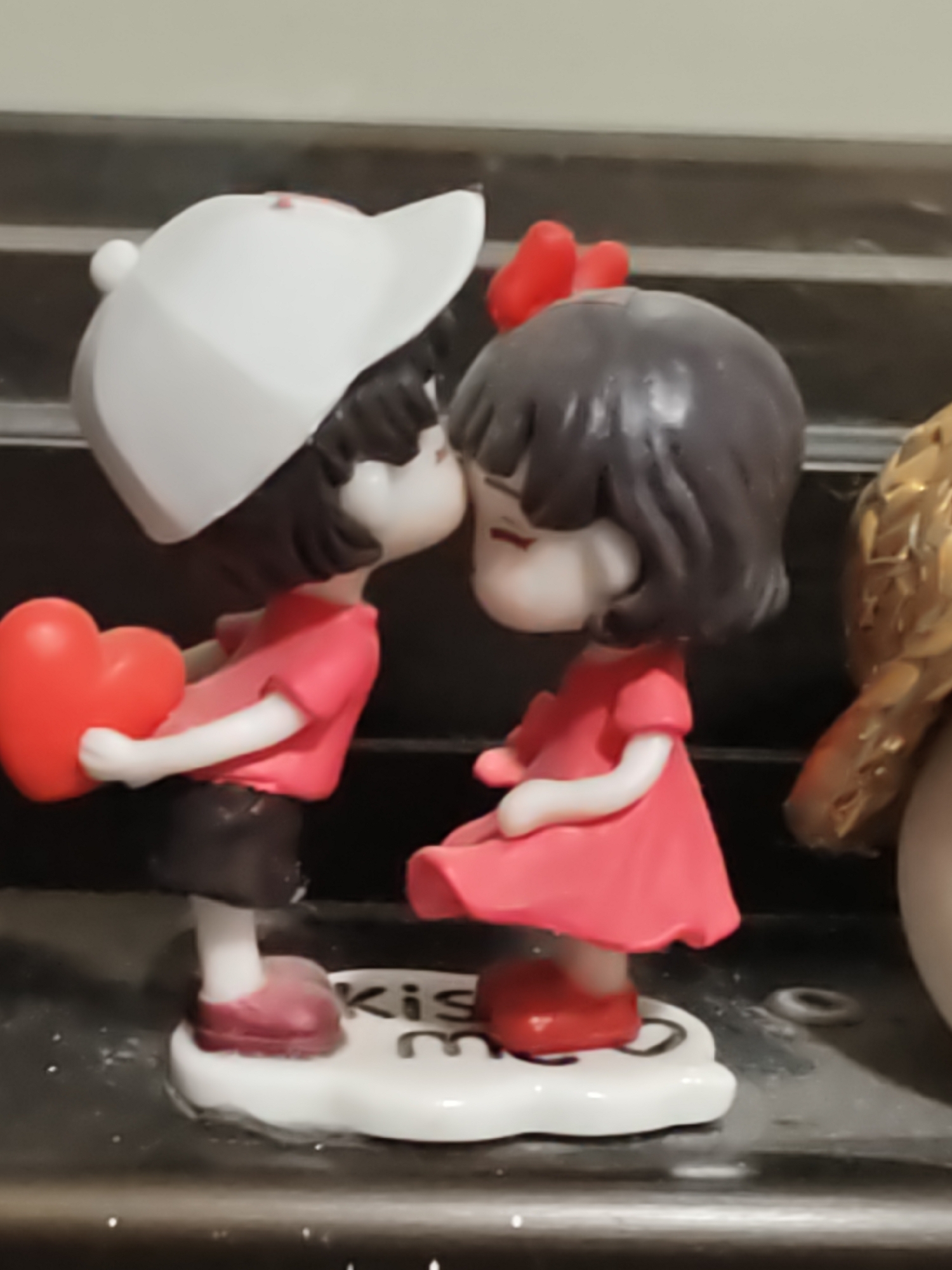 The image shows a close-up of two small figurines, a boy and a girl, depicted in a romantic embrace. The boy, on the left, is wearing a white baseball cap and a pink t-shirt, holding a red heart. The girl, on the right, is wearing a pink dress and has a red bow in her hair. Both figurines are on a white base that reads "Kiss me". The figurines are positioned on a dark surface, with a blurred background that suggests an indoor setting. There is no indication of the location being Petah Tikva, Israel.