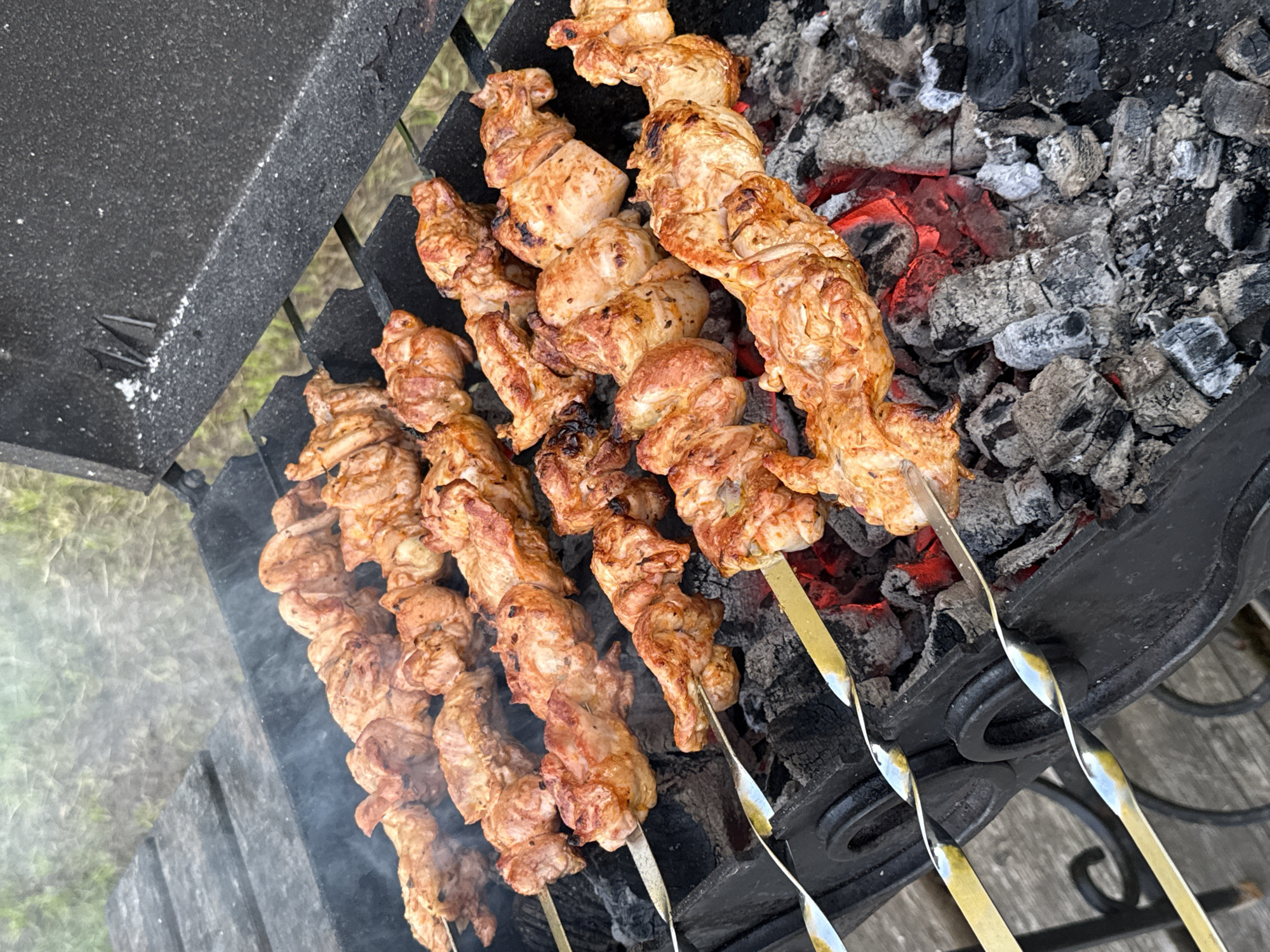 The image shows chicken skewers cooking on a charcoal grill. The grill is black metal, and the skewers are made of metal. The chicken is browned and appears to be partially cooked. Smoke is rising from the grill, indicating that it is in use. The background is blurred, but it appears to be an outdoor setting with some greenery. The location is identified as Zamosh'e (Zaklinskoe S/P), Russia. There are no people visible in the image.