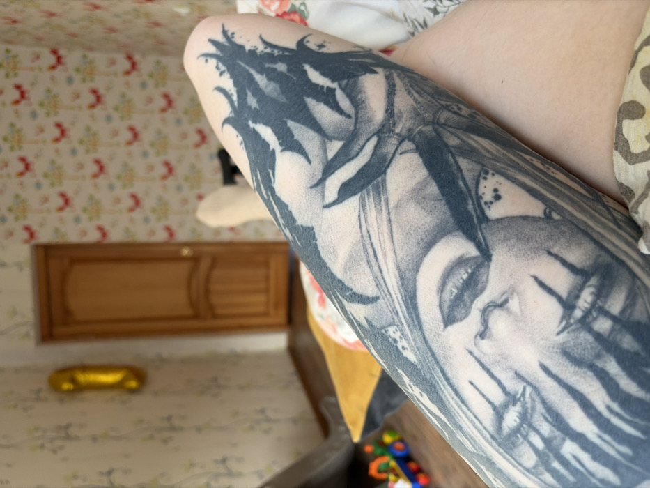 The image depicts an indoor, likely residential, scene in Chaikovsky, Russia, viewed from a first-person perspective. The primary subject in the foreground is a fair-skinned human leg, heavily tattooed with a large-scale black and grey design. The tattoo features a stylized face with closed or downward-looking eyes, prominent dark vertical markings across the cheeks, and is surrounded by dense, dark, organic or botanical elements. A light-colored fabric with a floral pattern is visible at the bottom right.
In the blurred background, another fair-skinned foot, potentially wearing a light sock, is visible to the upper right. A dark, indeterminate object, possibly a handgun or remote, is partially obscured behind this foot. To the left, a yellow cushion rests on a dark surface, and a multi-colored children's toy with wheels is on the floor. The walls are covered with wallpaper; the left wall features a muted pattern, while the right wall displays a repetitive pattern of red and yellow stylized motifs on a light background. A light brown wooden paneled door with a gold-colored doorknob is centrally located in the background. A golden, elongated, reflective balloon is affixed to the left wall. The floor appears to be dark wood or laminate.