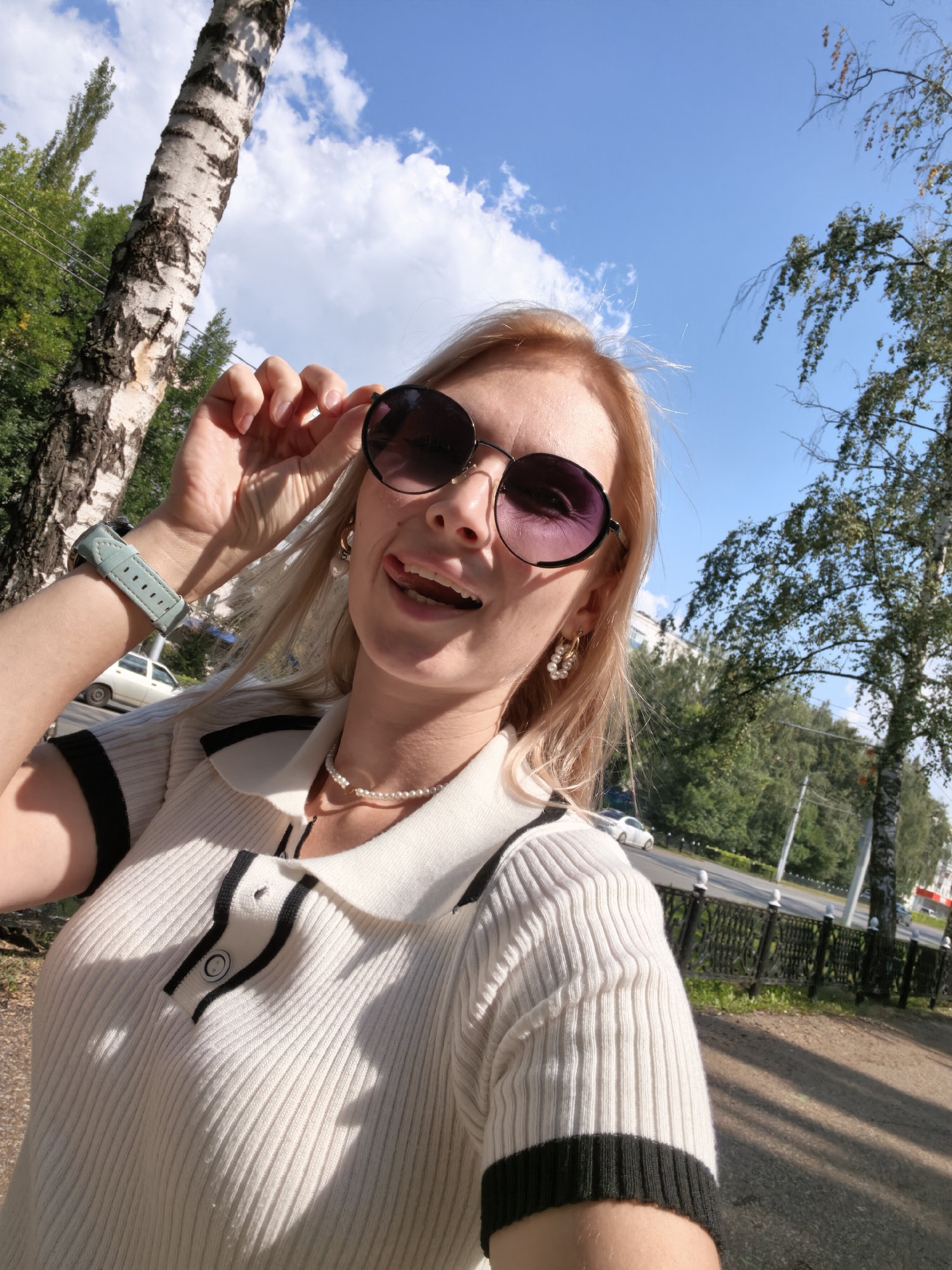 A young woman is taking a selfie outdoors in Sterlitamak, Russia. She is wearing sunglasses, a white ribbed polo shirt with black trim, and a pearl necklace. She is sticking her tongue out at the camera. In the background, there is a birch tree, green foliage, a street with cars, and a blue sky with white clouds.