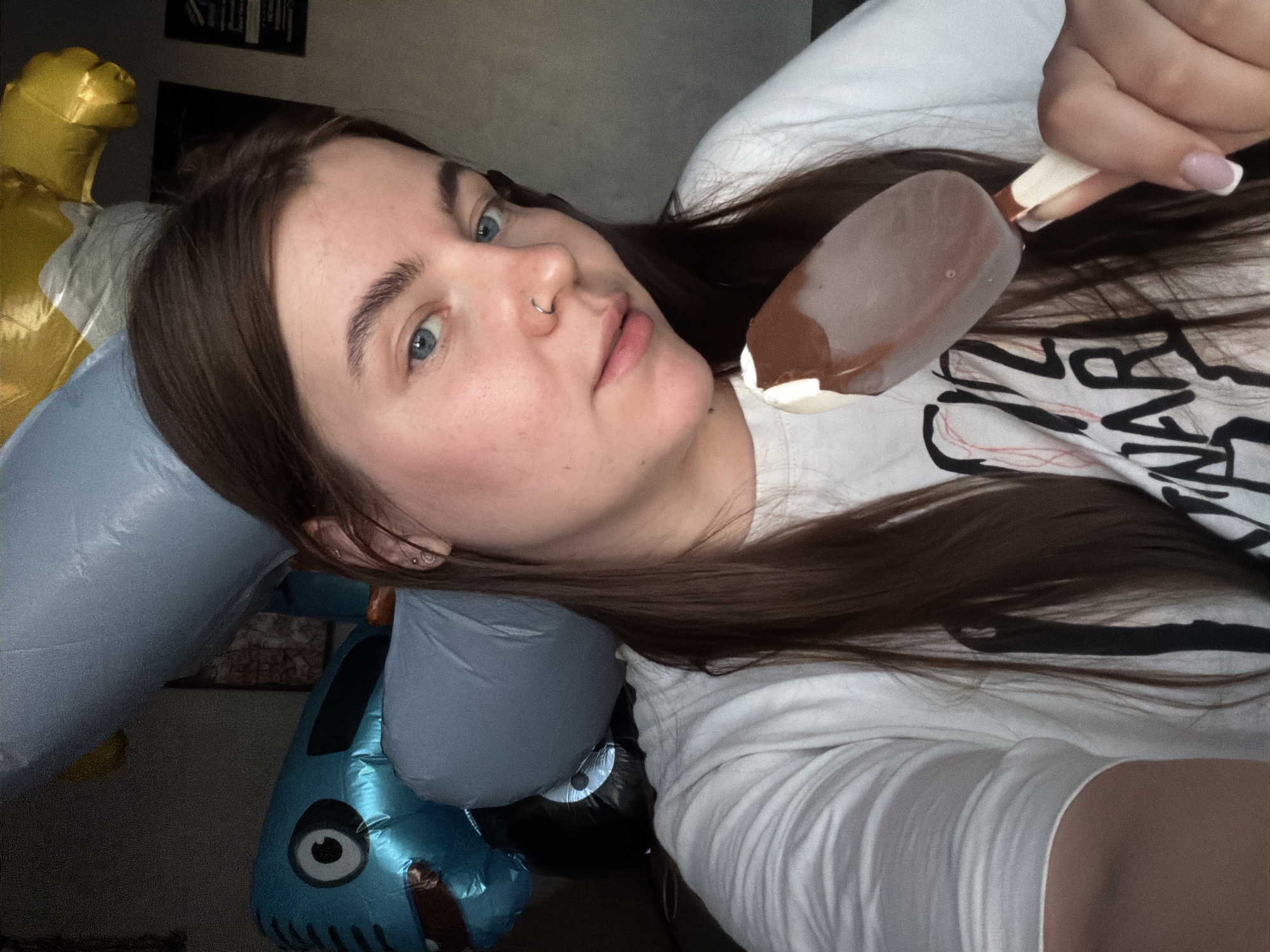 A young woman with long brown hair and blue eyes is taking a selfie while holding a chocolate-covered ice cream bar. She is wearing a white t-shirt with black lettering. In the background, there are several inflatable balloons, including a blue cartoonish character. The scene appears to be indoors, likely a residential setting. No specific location in Chaikovsky, Russia can be determined from the visual information provided.