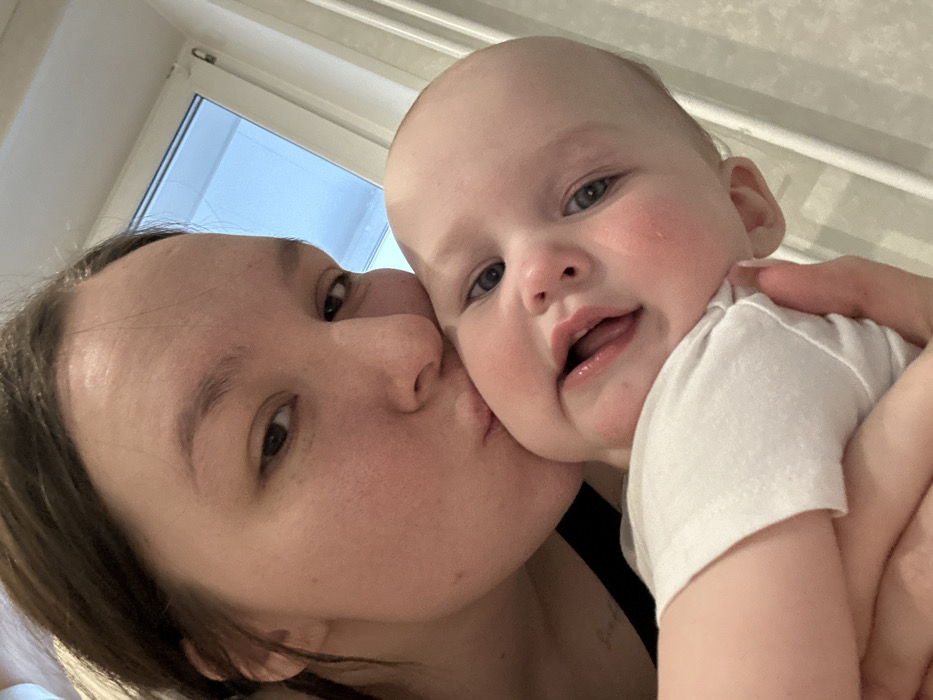 The image presents a close-up indoor view of an adult female kissing an infant. The female has brown hair and is seen with her right eye and nose visible, lips pursed against the infant's left cheek. She wears a dark top, with faint markings visible on her upper chest area. The infant, wearing a white short-sleeved top, has light eyes and a slightly open mouth, looking towards the viewer. The background features a light-colored wall and a white-framed window in the upper right quadrant, displaying a light blue exterior. No visual identifiers are present to specify a precise location within Chaikovsky, Russia.