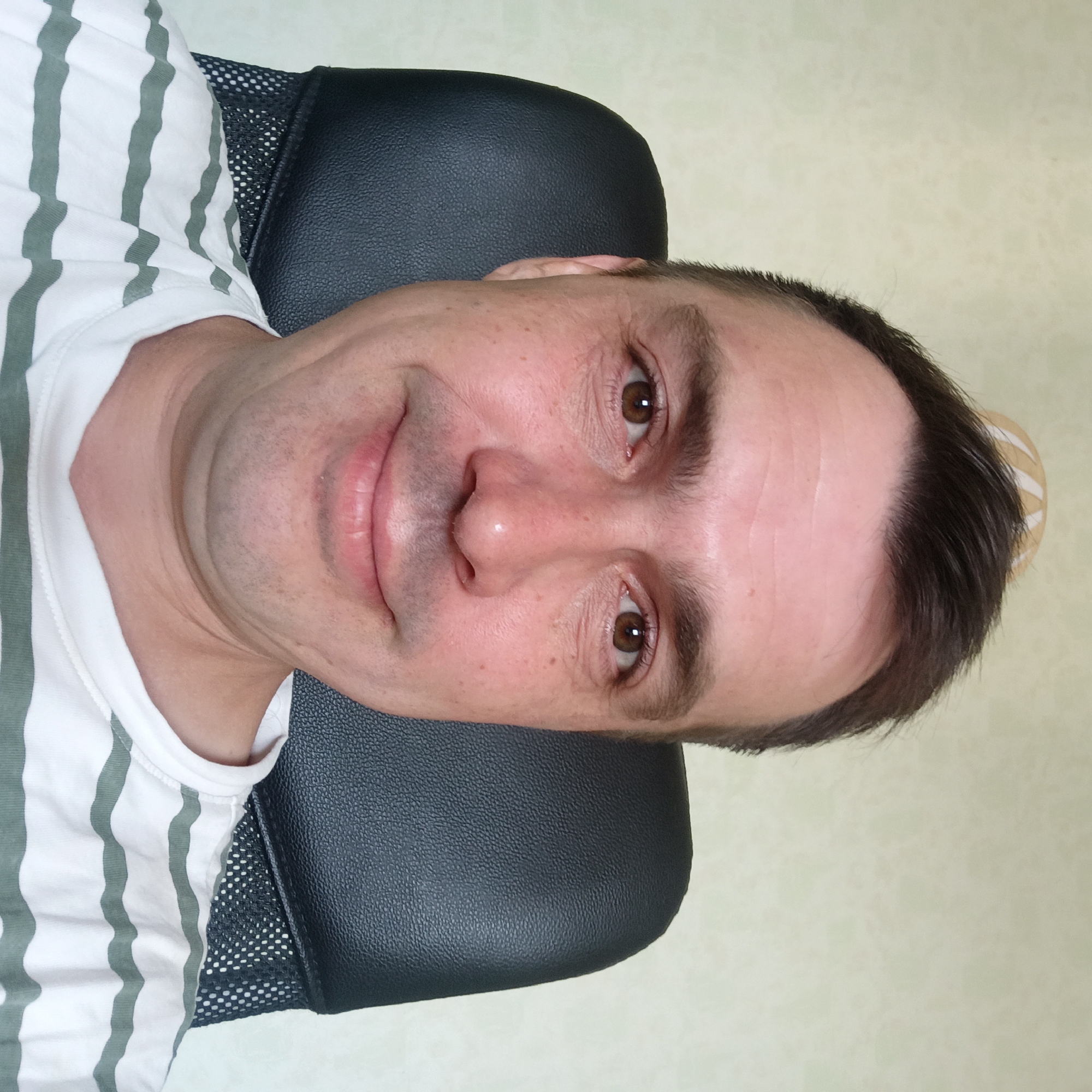 A medium shot shows a man from the chest up, sitting in a black office chair. He is looking directly at the camera with a slight smile. The man has short, dark brown hair, brown eyes, and fair skin with some freckles. He is wearing a white t-shirt with thin green stripes. The background is a plain, light-colored wall, possibly wallpapered. The scene appears to be indoors, in a domestic or office setting. There is no identifiable signage or context to place the image in Nizhny Novgorod, Russia.