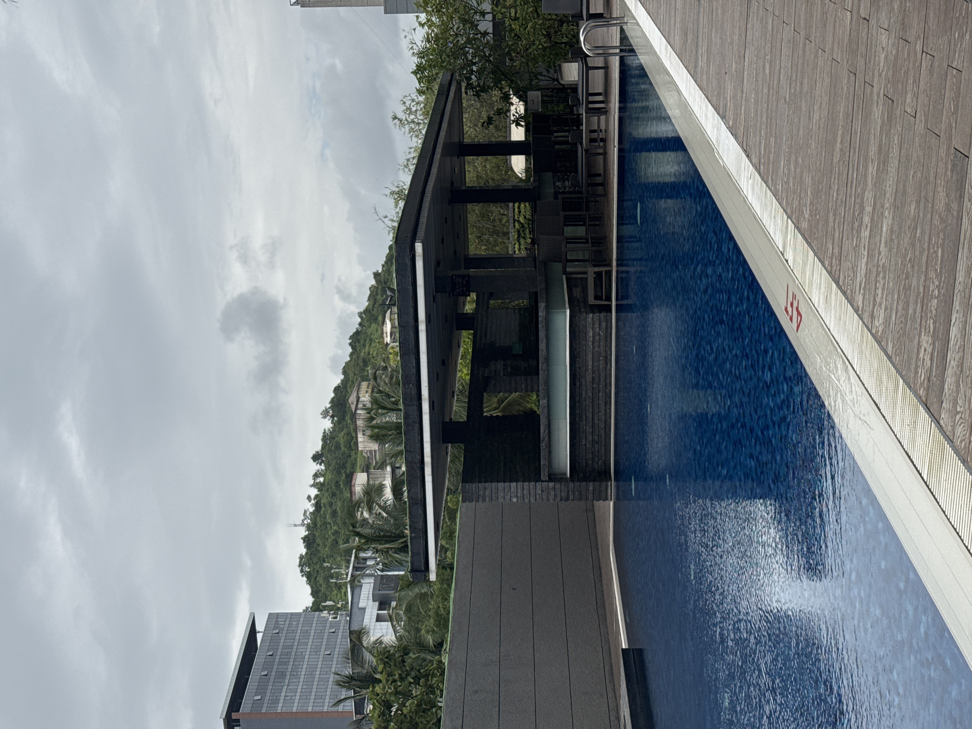 The image shows a swimming pool area at a resort or hotel, likely located in Pune, India, given the cloudy, overcast sky and lush green hills in the background. In the foreground, a portion of the blue tiled swimming pool is visible, with a wooden deck bordering one side. The deck has a depth marker on the edge of the pool indicating "4 FT". Beyond the pool, there is a modern open-air bar with a dark roof supported by pillars, furnished with bar stools and tables. A dark gray textured wall runs behind the pool, separating it from the elevated area. In the background, a tall, multi-story building with many windows, lush green trees, and residential buildings are visible on a hillside under a cloudy sky. There are no people or animals visible in the scene. The overall atmosphere is one of a tranquil, possibly off-season, resort setting.